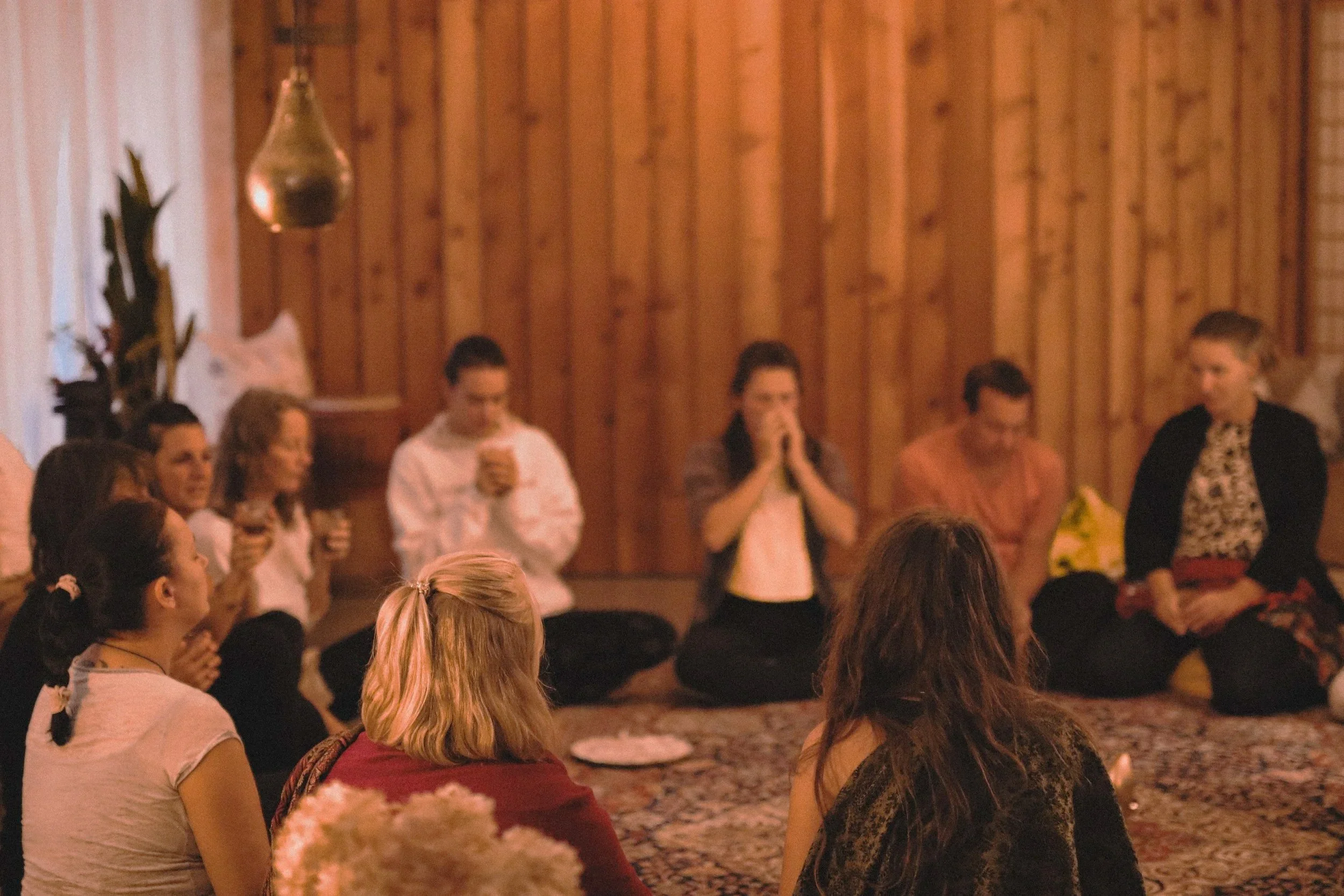 Eine Gruppe von Menschen sitzt auf einem Teppich in einem Raum mit Holzvertäfelung. Meditationskreis mit warmer Beleuchtung und entspannter Atmosphäre.