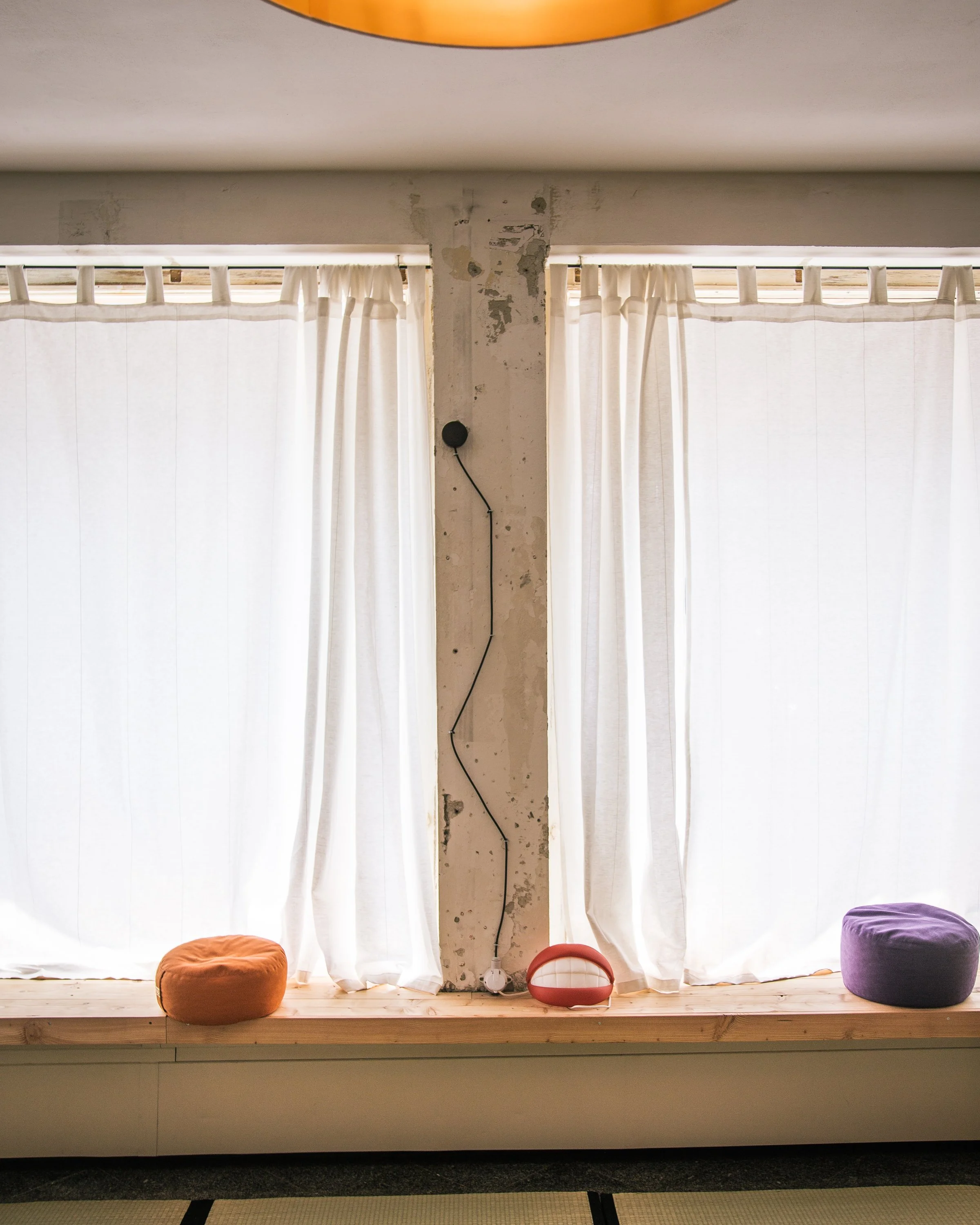 Ein Fenster mit weißen Vorhängen, auf einem Holzfensterbrett stehen drei runde Sitzkissen in Orange, Weiß und Lila. Dazwischen steht eine Lampe in Lippenform.