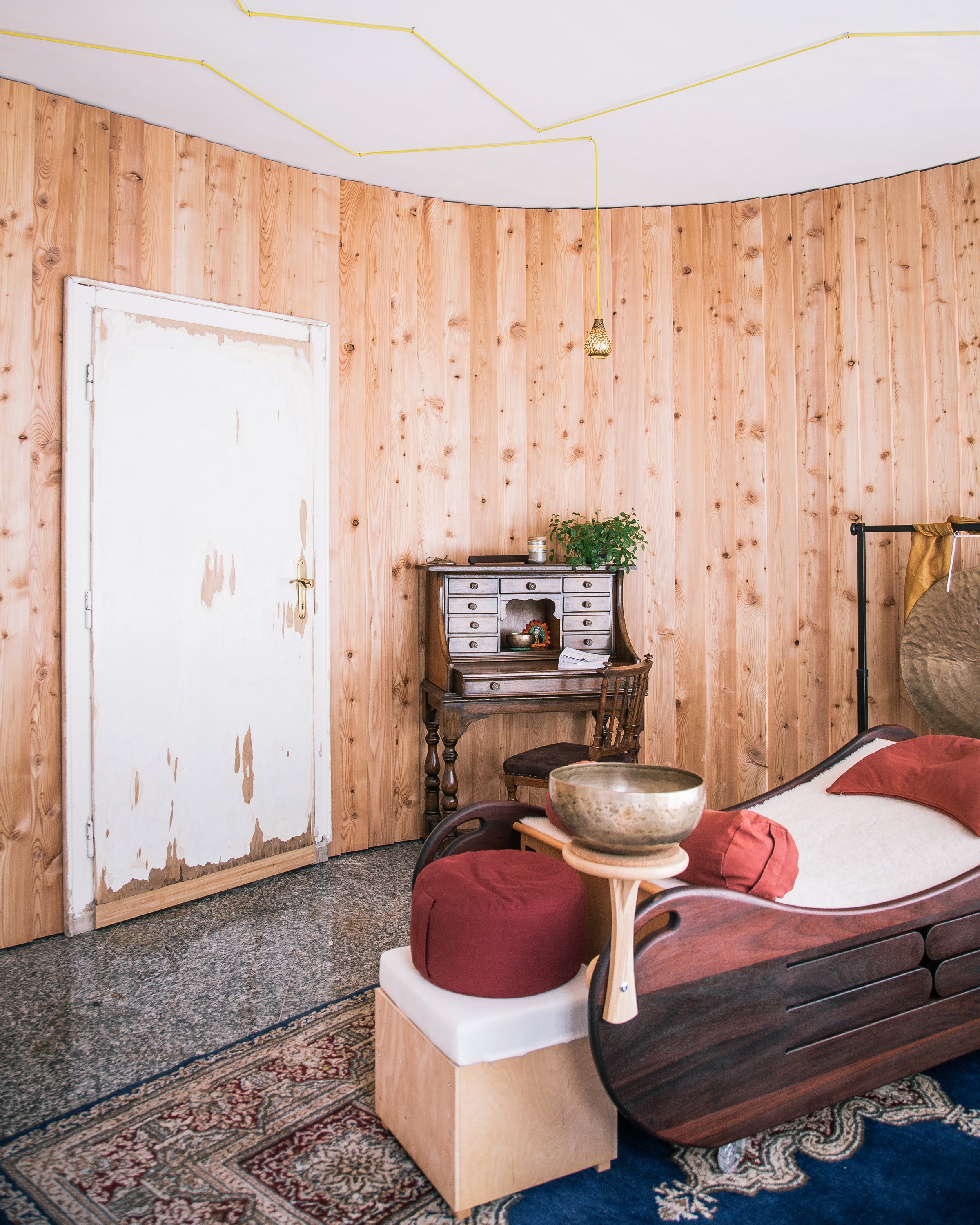 Ein gemütlicher Praxisraum mit Holzvertäfelung, antiker Schreibkommode, Klangbett mit roten Kissen, einer kleinen Sitzbank,.Der Boden ist mit einem Teppich bedeckt.