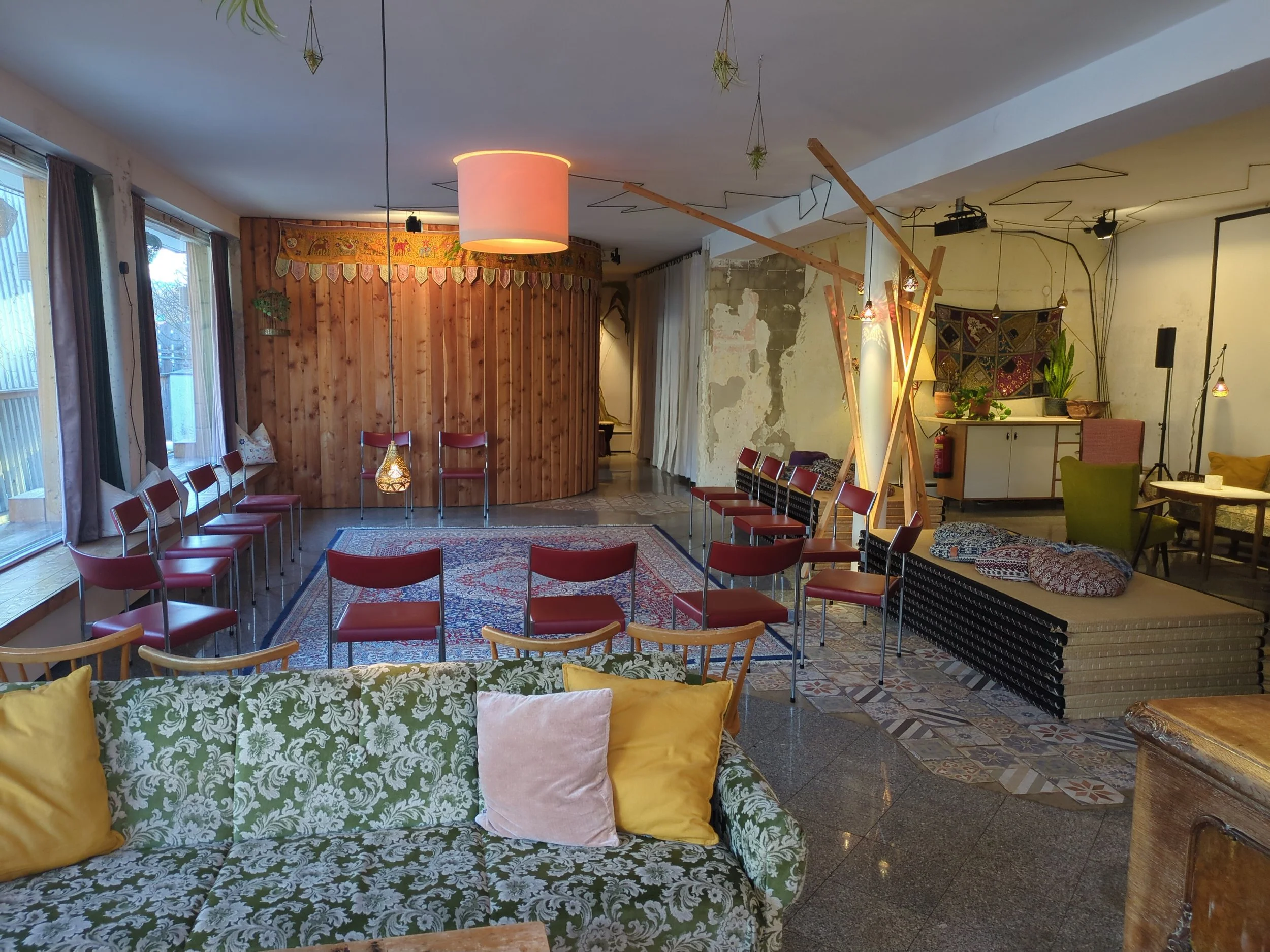 Innenansicht eines gemütlichen Raumes mit Sessel, Stühlen, Teppichen und Dekorationen, ein Bereich mit Säulen und Hängepflanzen.