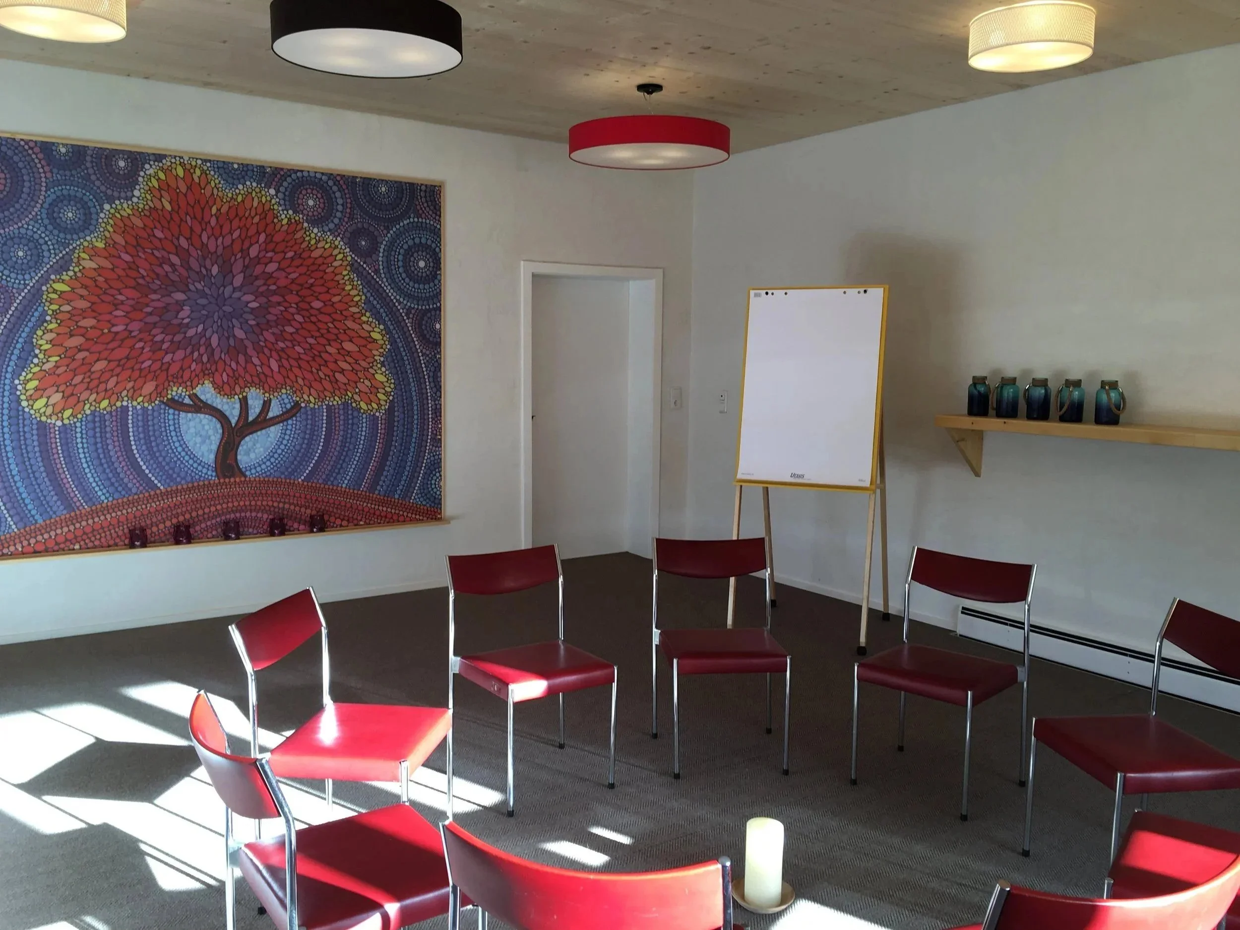 Ein Sitzungsraum mit mehreren roten Stühlen, einem weißen Flipchart, einer Wand mit einem bunten Baum-Bild und einer Holzregal mit blauen Kerzenhaltern.