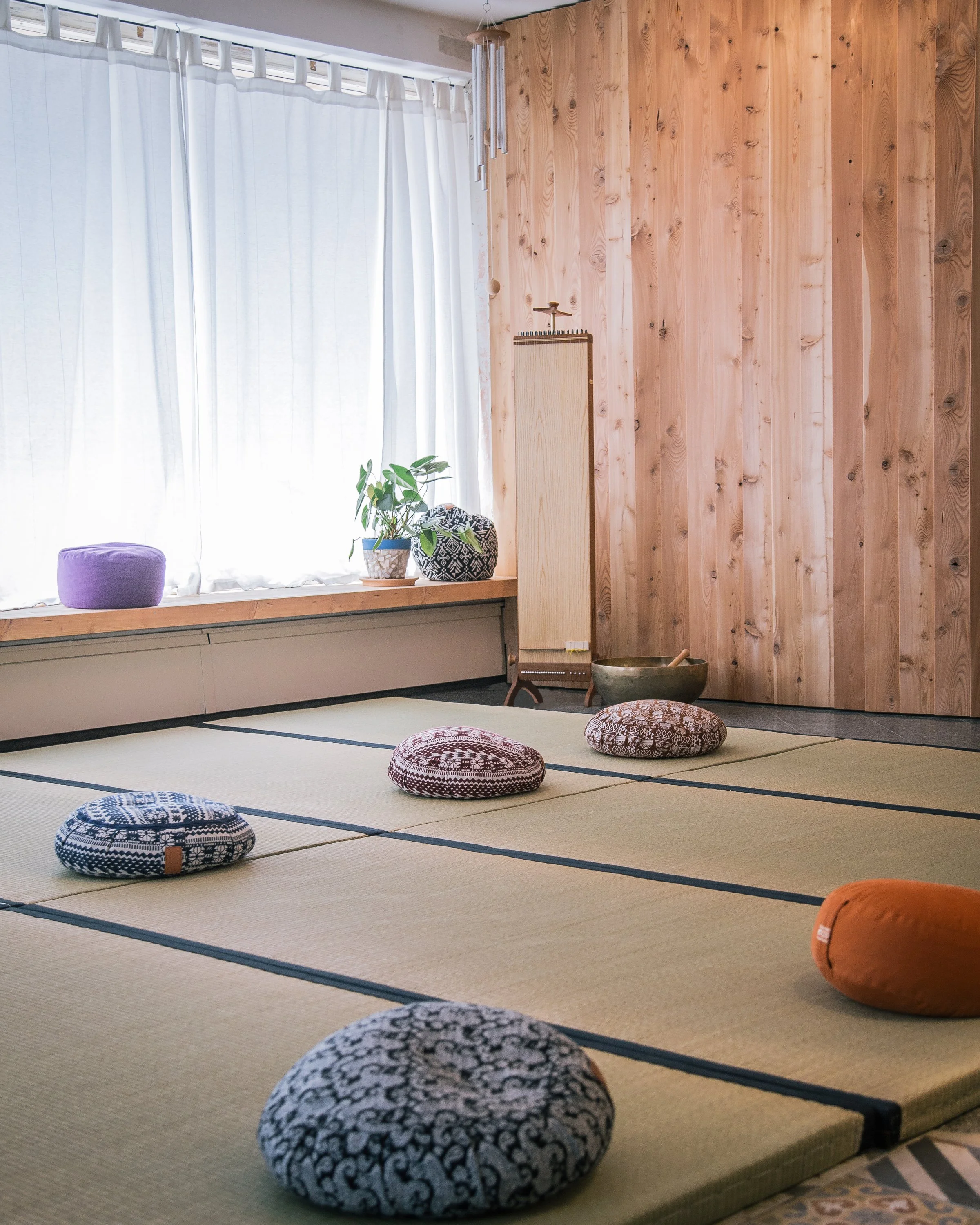 Ein Raum mit Tatami-Matten, runden Kissen, einer Wand aus Holzpanelen, einer großen Fensterfront mit weißen Vorhängen, Pflanzen auf einem Fensterbrett, einem Windspiel an der Decke, einer Klangschale und einem Raumteiler.