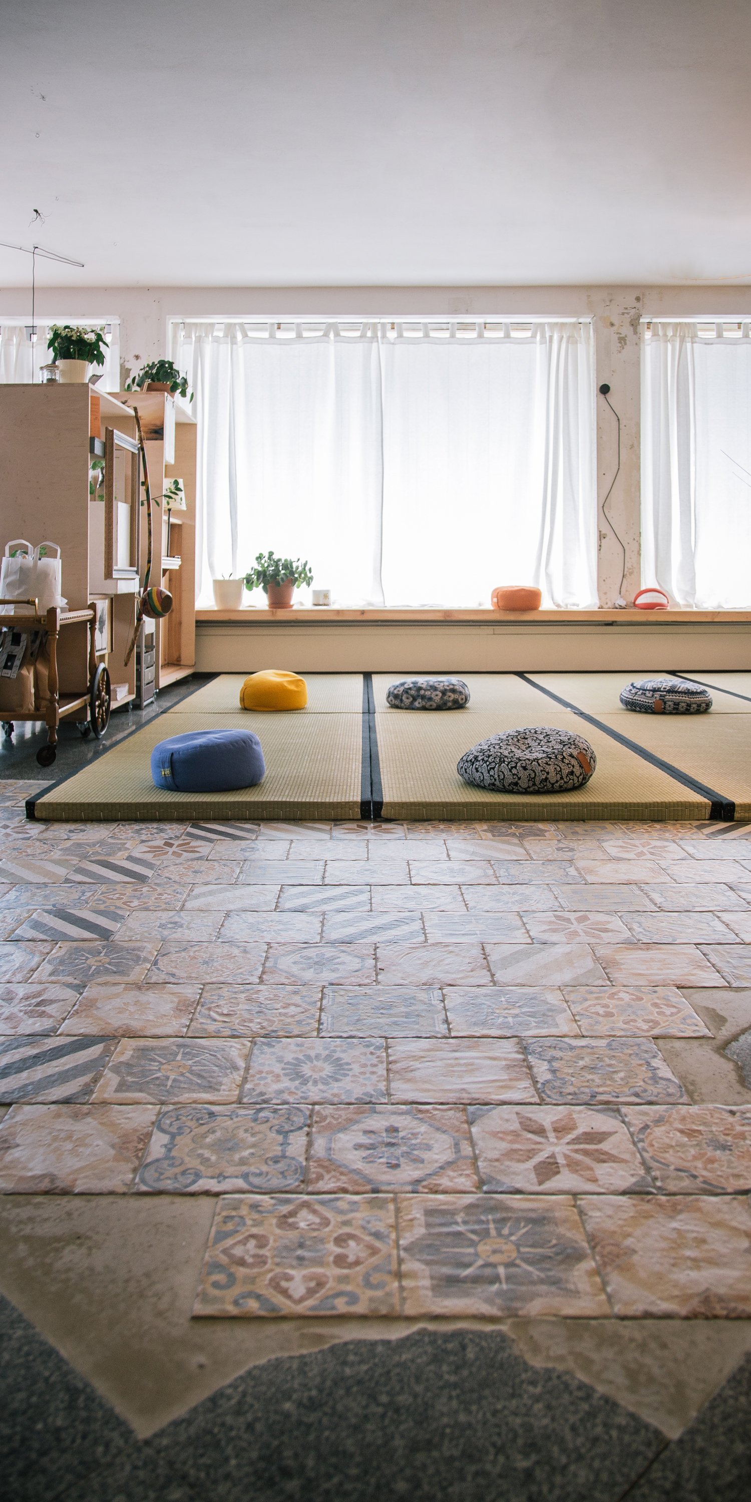 Ein Raum mit Tatami-Matten und Kissen, helle Vorhänge vor großen Fenstern, der Raum wirkt wohnlich und ruhig.