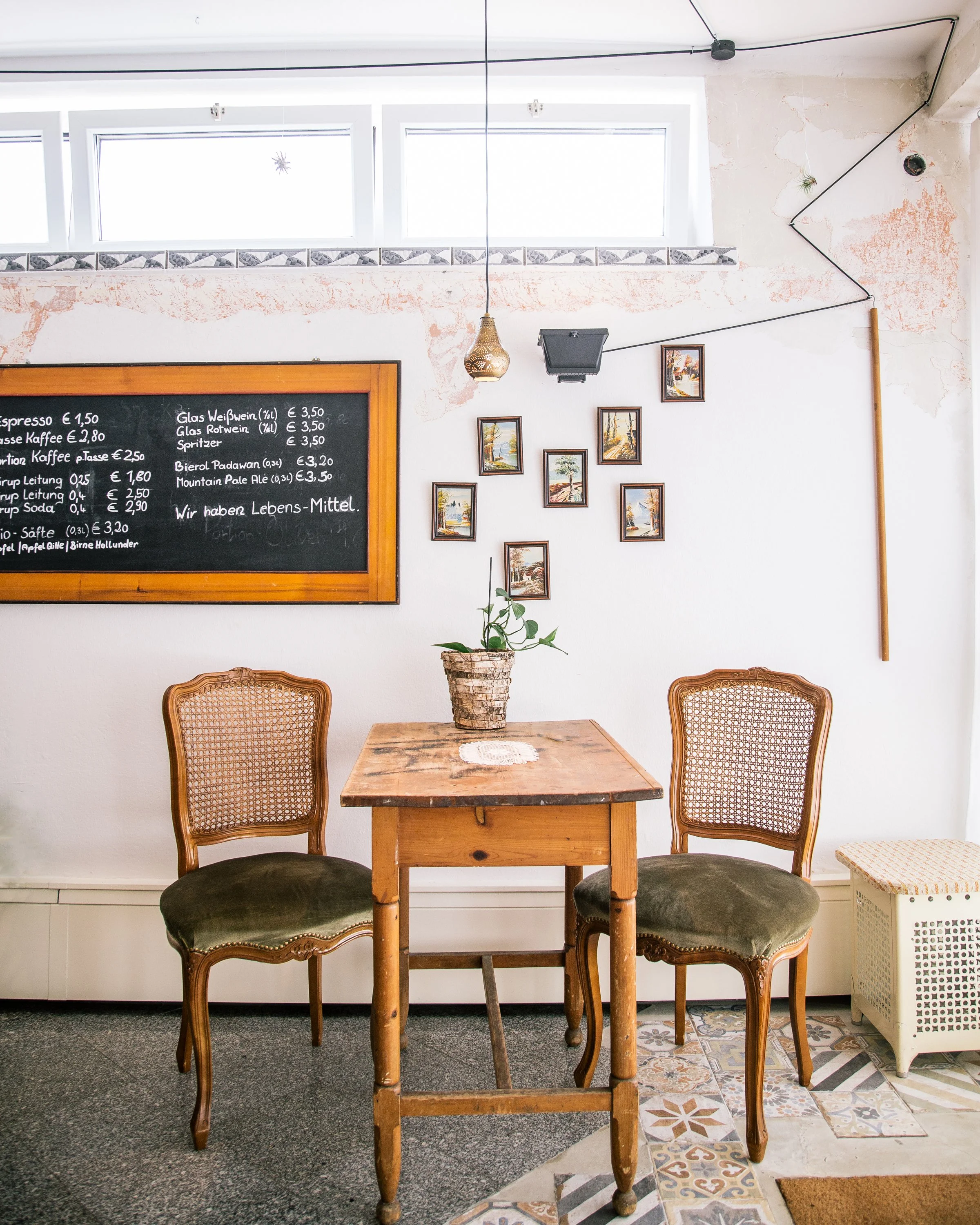 Ein kleiner Café-Tisch mit zwei alten, gepolsterten Stühlen vor einer weißen Wand. Auf dem Tisch steht eine Topfpflanze. An der Wand hängt eine schwarze Kreidetafel. Es gibt eine Deckenleuchte und eine dekorative Wandmontage.