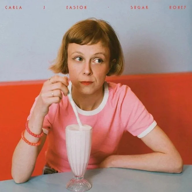 Carla J Easton Album artwork - I Think That I Might LA woman with short, light brown hair, wearing a pink t-shirt, sitting at a table and stirring a milkshake with a straw. The background has the words 'Carla J Fashion Sugar Honey' written across it.
