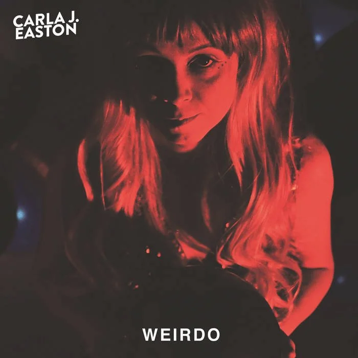 Carla J Easton Album artwork - I Think That I Might Love You. A woman with long, wavy blonde hair illuminated by red light, smiling at the camera. Text on the image includes "Carla J. Easton" in the top left corner and "Weirdo" at the bottom.