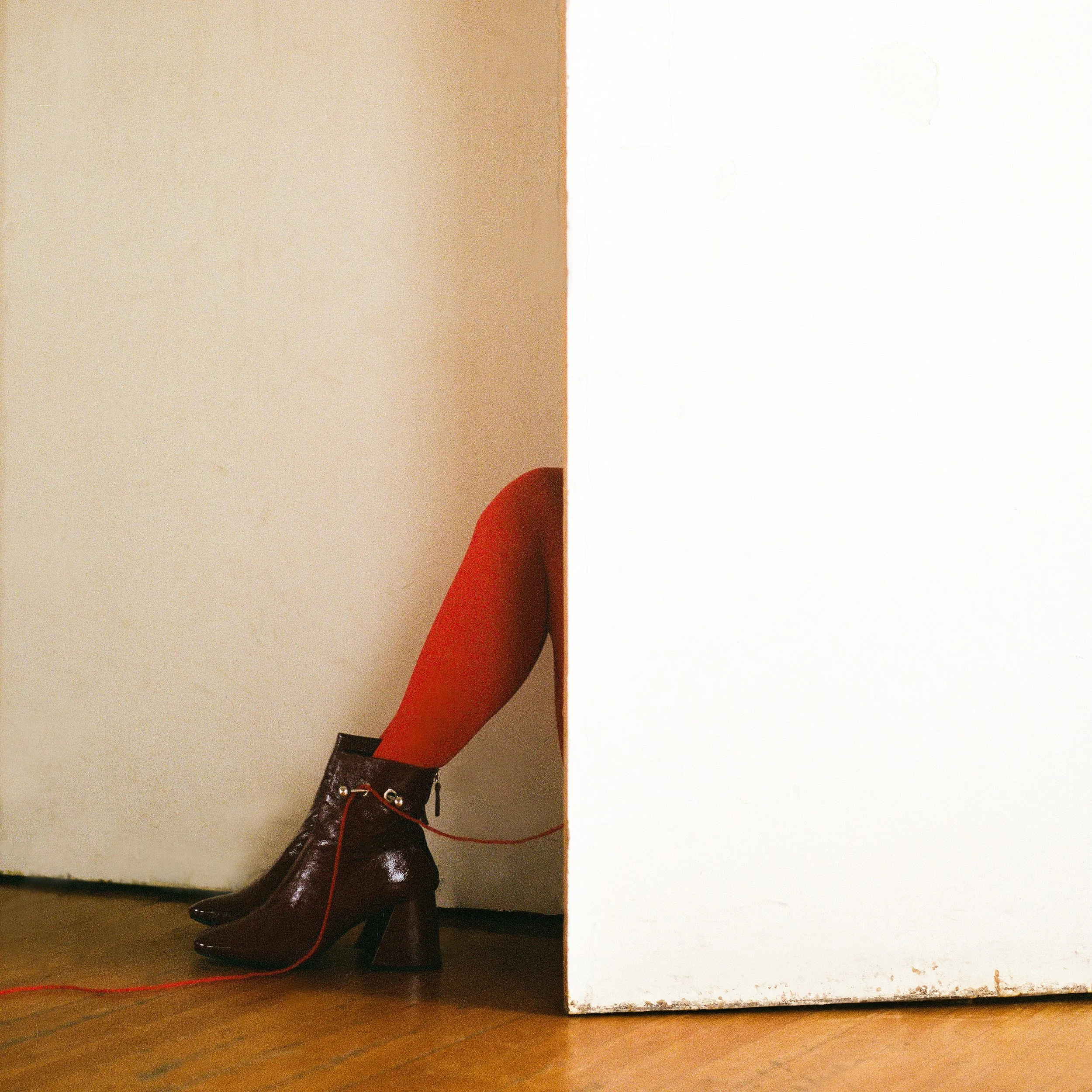 Carla J Easton Album artwork - I Think That I Might Love You A person wearing red tights and shiny black ankle boots with a chunky heel is partially hidden behind a white wall, with only their leg visible.