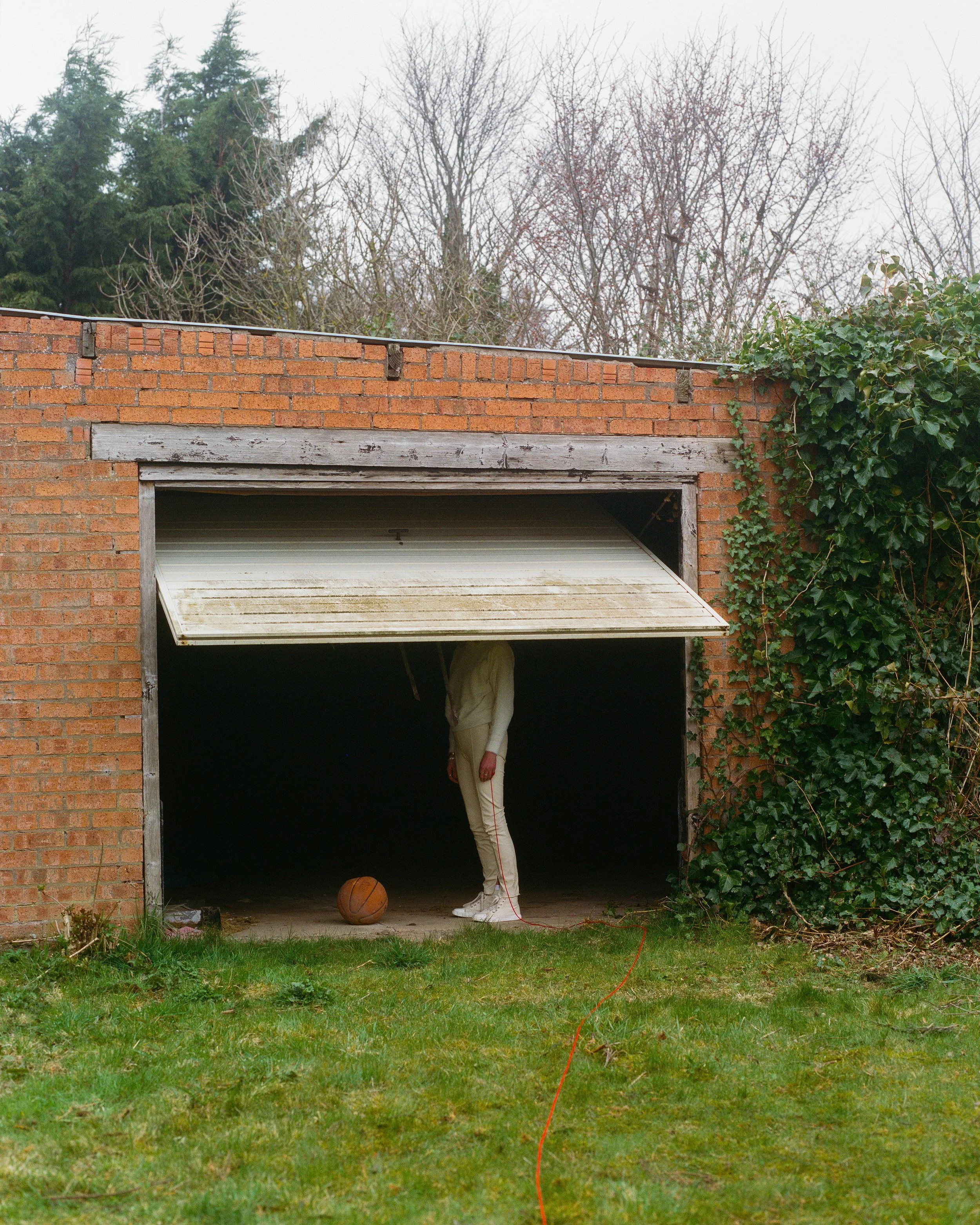 Carla J Easton album artwork. A person dressed in white standing in a dark garage with a basketball on the ground and an orange extension cord trailing outside, surrounded by greenery and a brick wall.