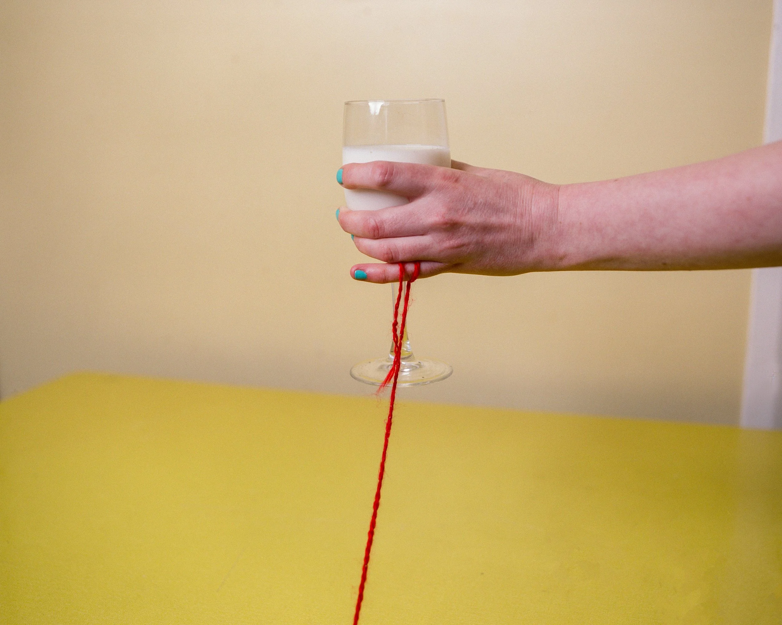 Carla J Easton Album artwork - I Think That I Might Love You A hand with turquoise painted nails holding a stemmed glass of milk, with a red string hanging from the base of the glass, against a yellow background.