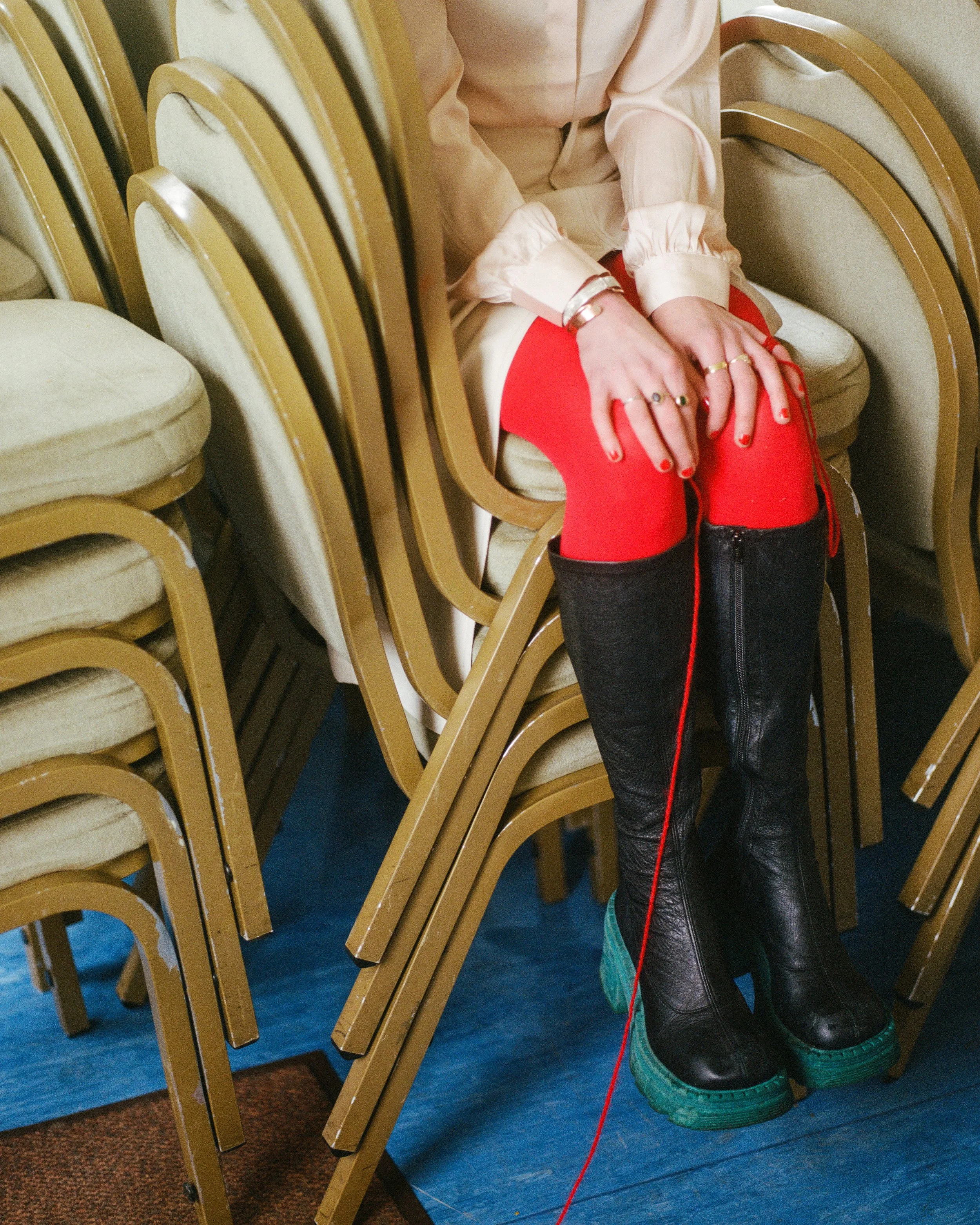 Carla J Easton album artwork. A person sitting on stacked chairs, wearing a beige shirt, red tights, and black platform boots with teal soles. They have jewelry on their hands and are surrounded by beige stacked chairs.