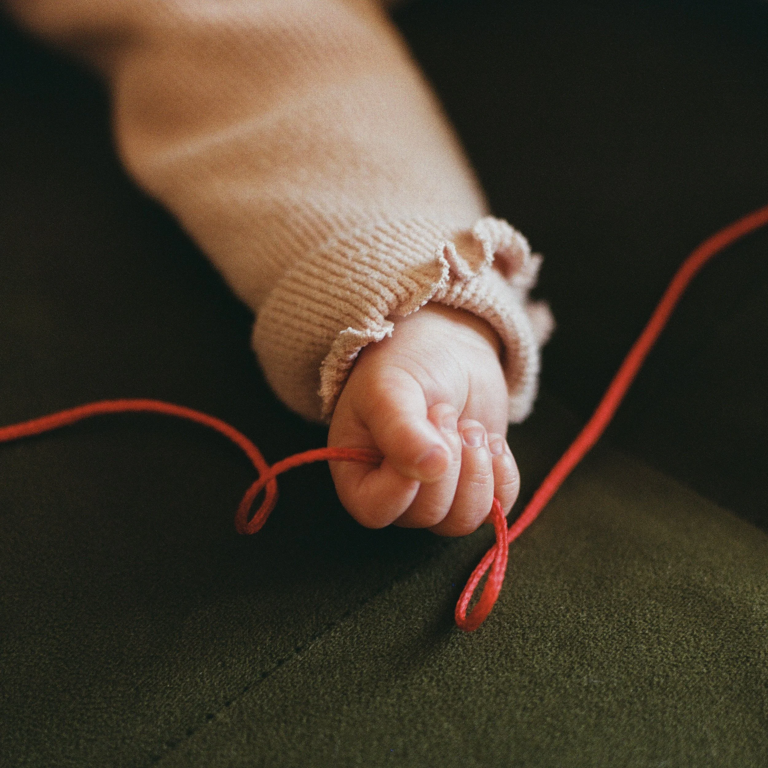 Carla J Easton Album artwork - I Think That I Might Love You Close-up of a baby's small hand holding a red shoelace, wearing a beige sweater with ruffled cuffs, resting on a dark surface.