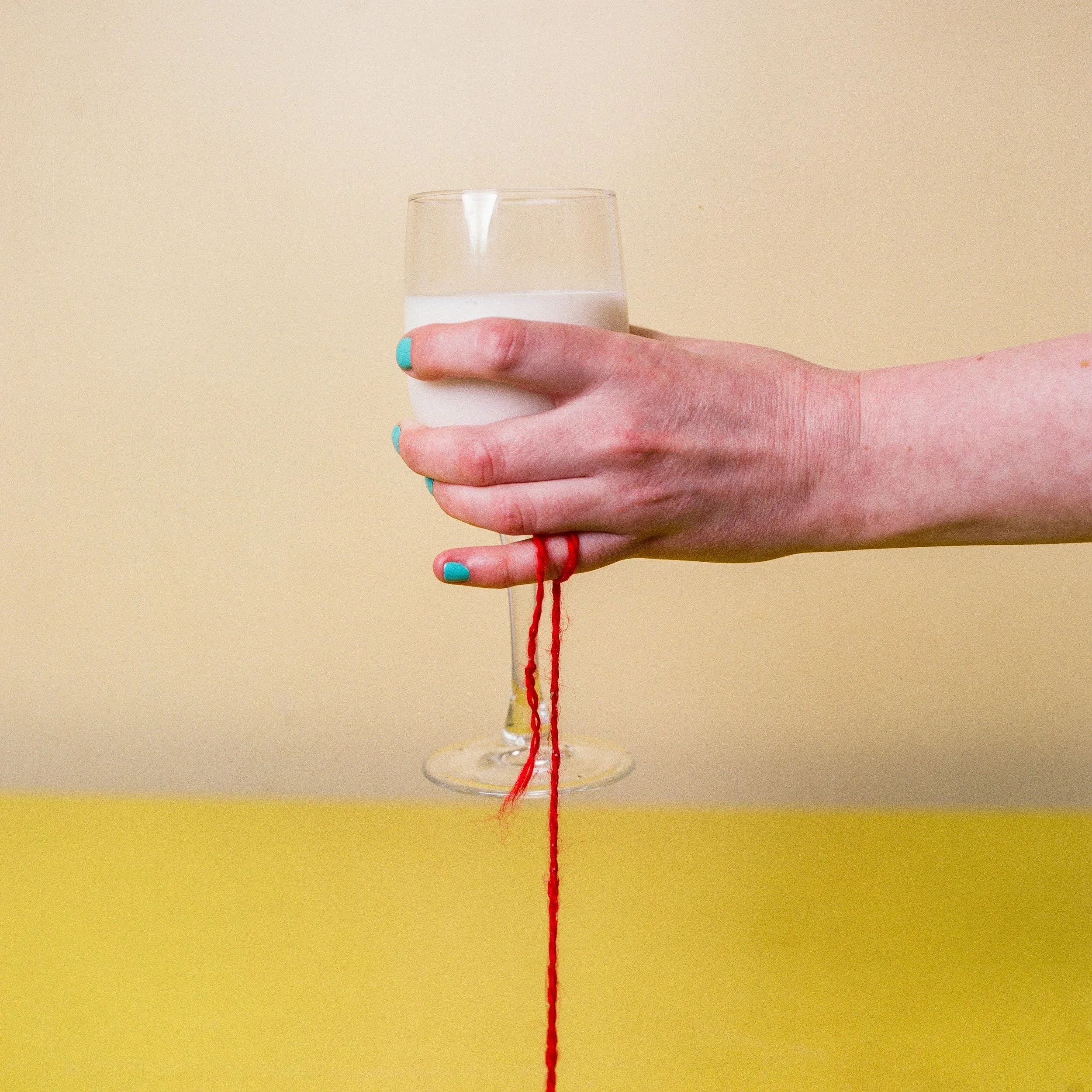 Carla J Easton Album artwork - I Think That I Might Love You A person's hand with turquoise-painted nails holding a glass of milk against a beige wall background, with a red thread hanging from the hand to a yellow surface.