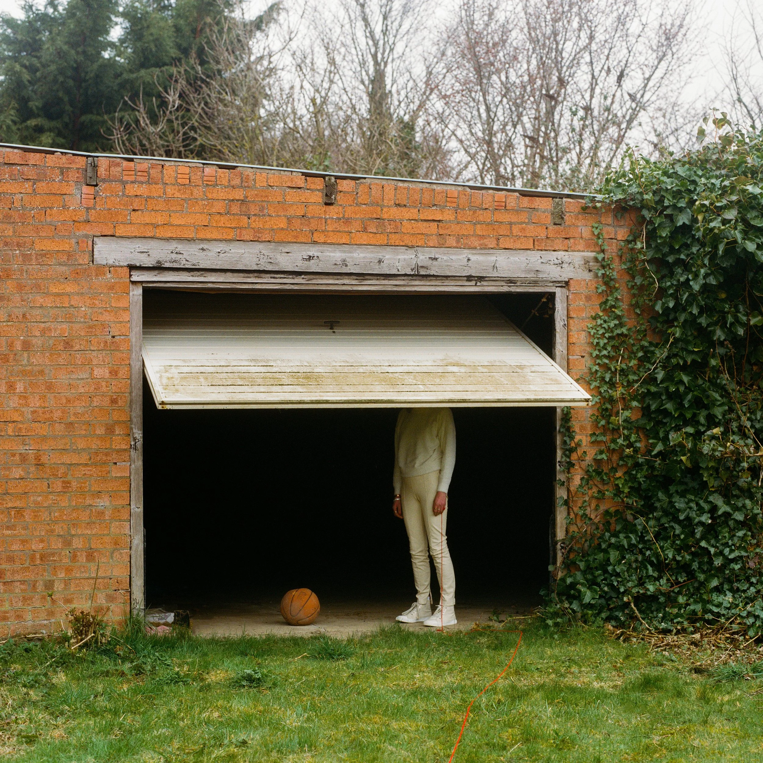 Carla J Easton Album artwork - I Think That I Might Love You Person in beige clothing standing in a dark garage with an open door, next to a basketball on the ground, and a grassy yard in front.