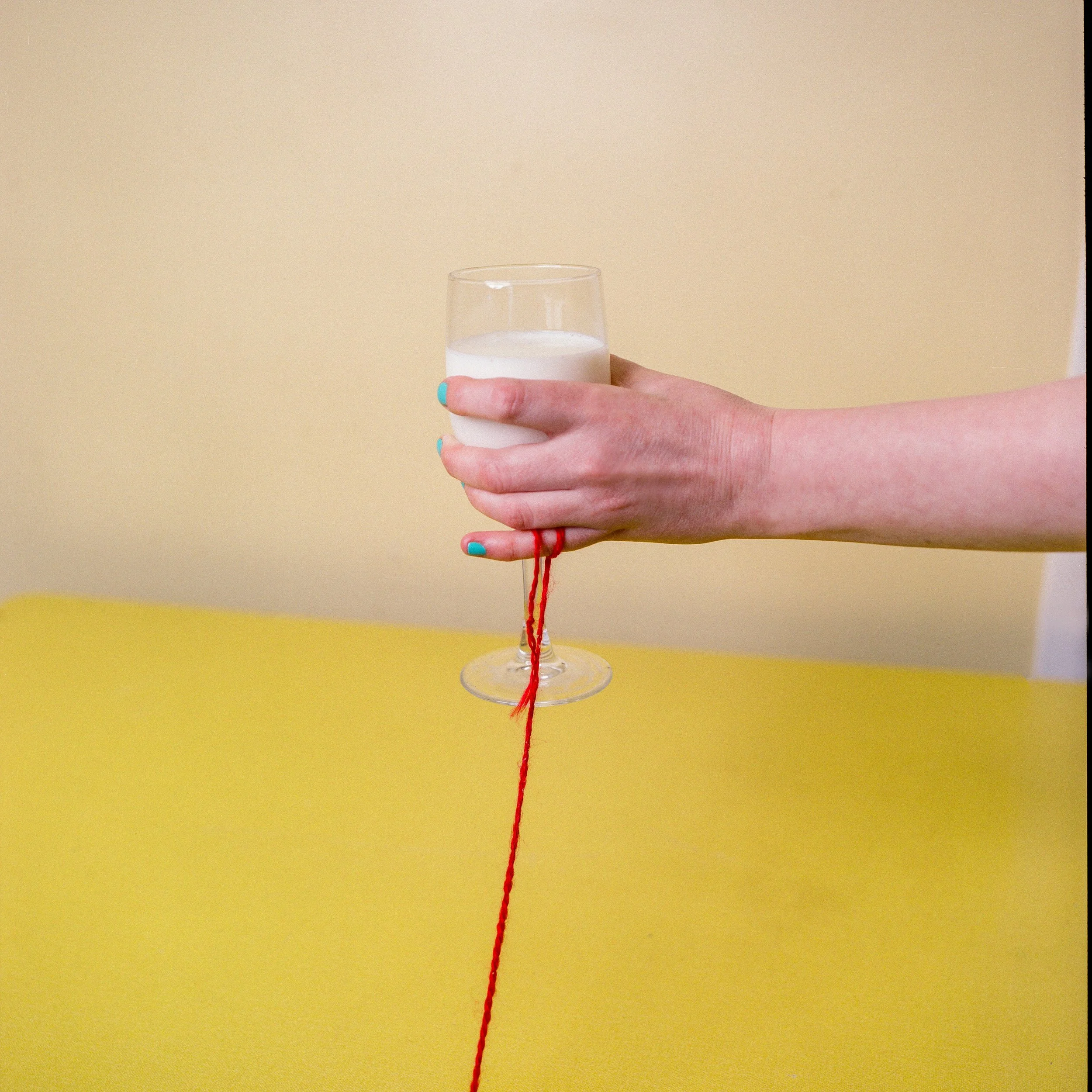 Carla J Easton Album artwork - I Think That I Might Love You Person holding a glass of milk with a red string attached to the bottom of the glass, against a yellow table and beige wall background.