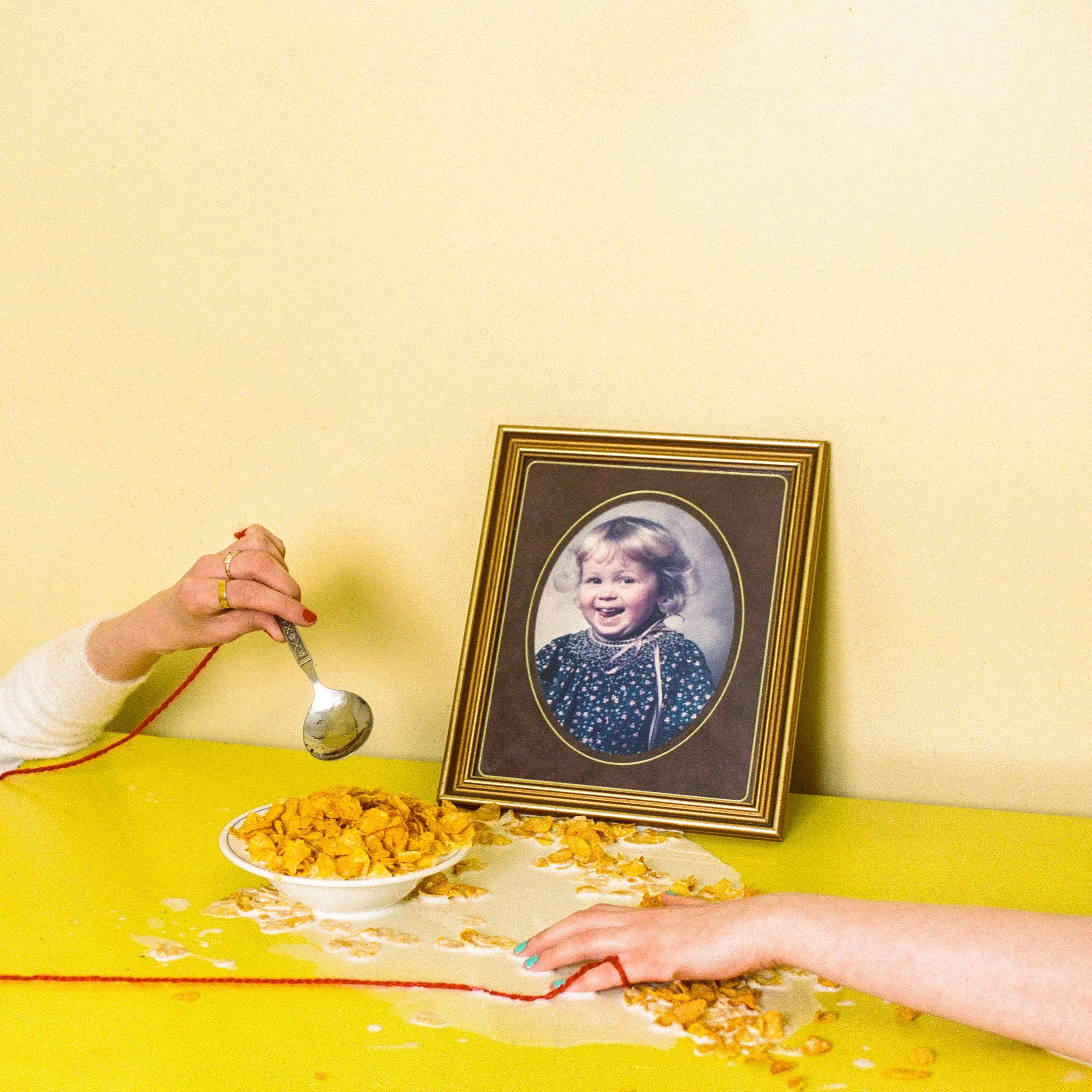 Carla J Easton Album artwork - I Think That I Might Love You A hand holding a spoon over a bowl of cereal with milk and spilled cereal on a yellow table, with a framed photograph of a smiling young girl on the table.
