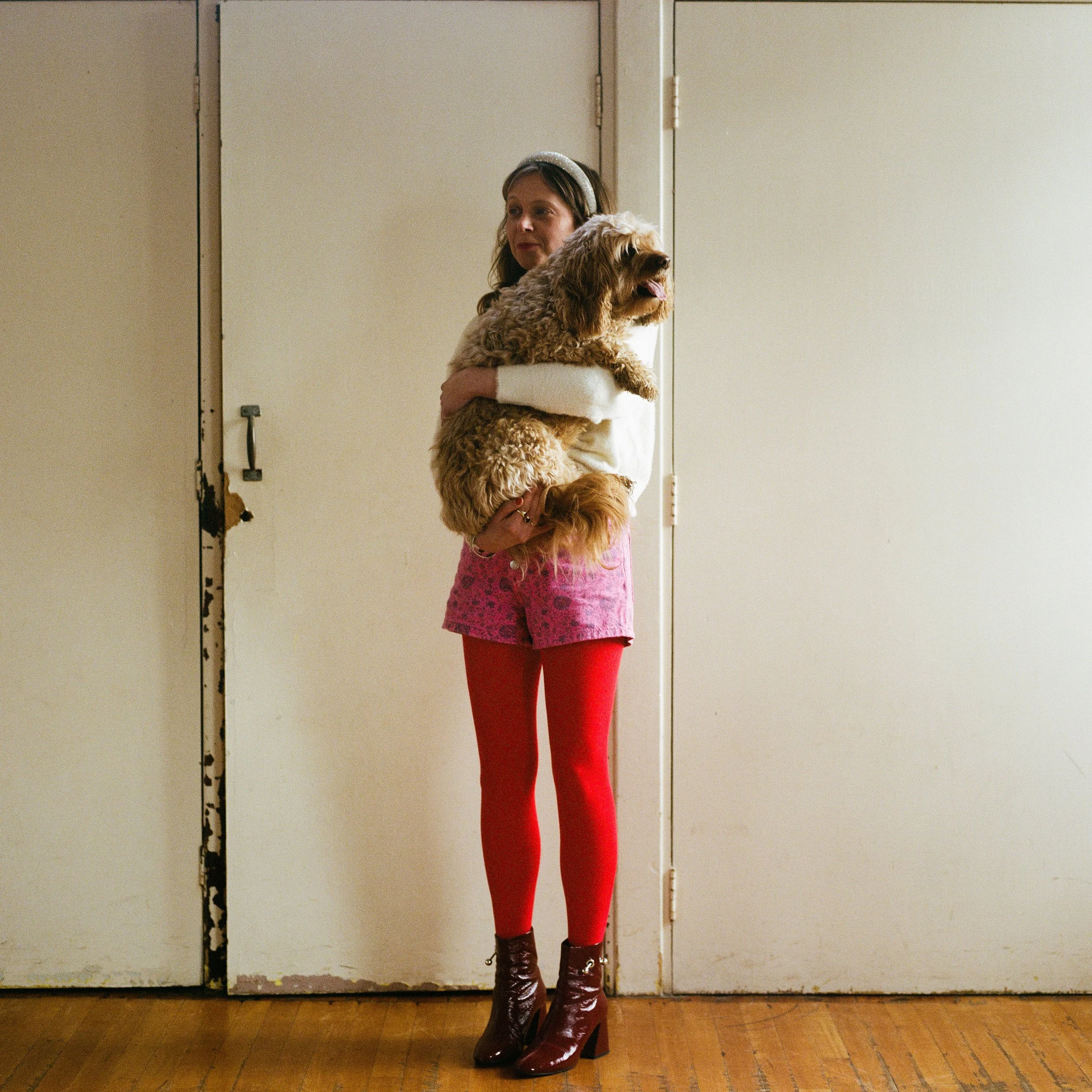 Carla J Easton Album artwork - I Think That I Might Love You A woman standing indoors holding a dog, wearing colorful clothing including red tights, pink shorts, a white sweater, and brown ankle boots, with a door and wooden floor in the background.