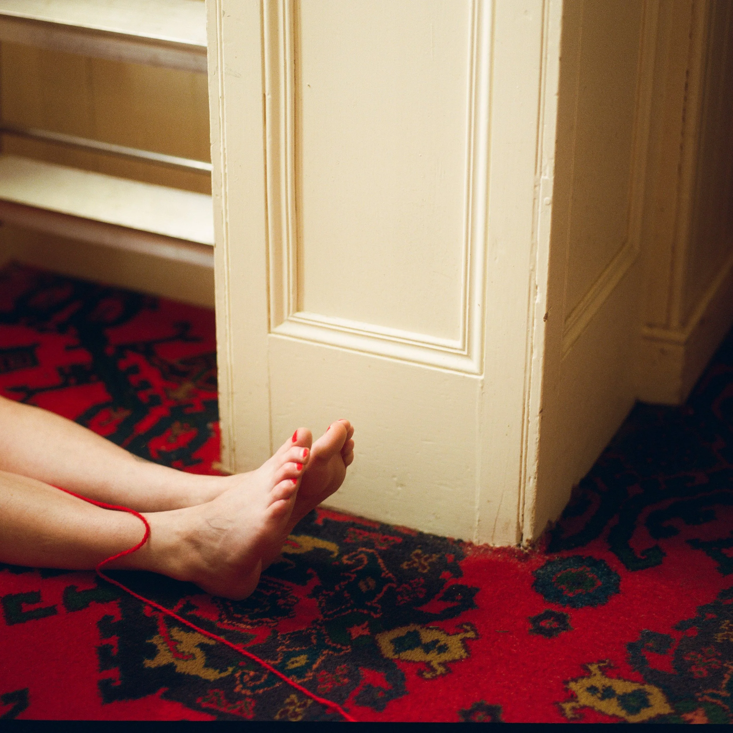 Carla J Easton Album artwork - I Think That I Might Love You Pair of bare feet with red-painted toenails and red string bracelet resting on a colorful patterned carpet near a white door.