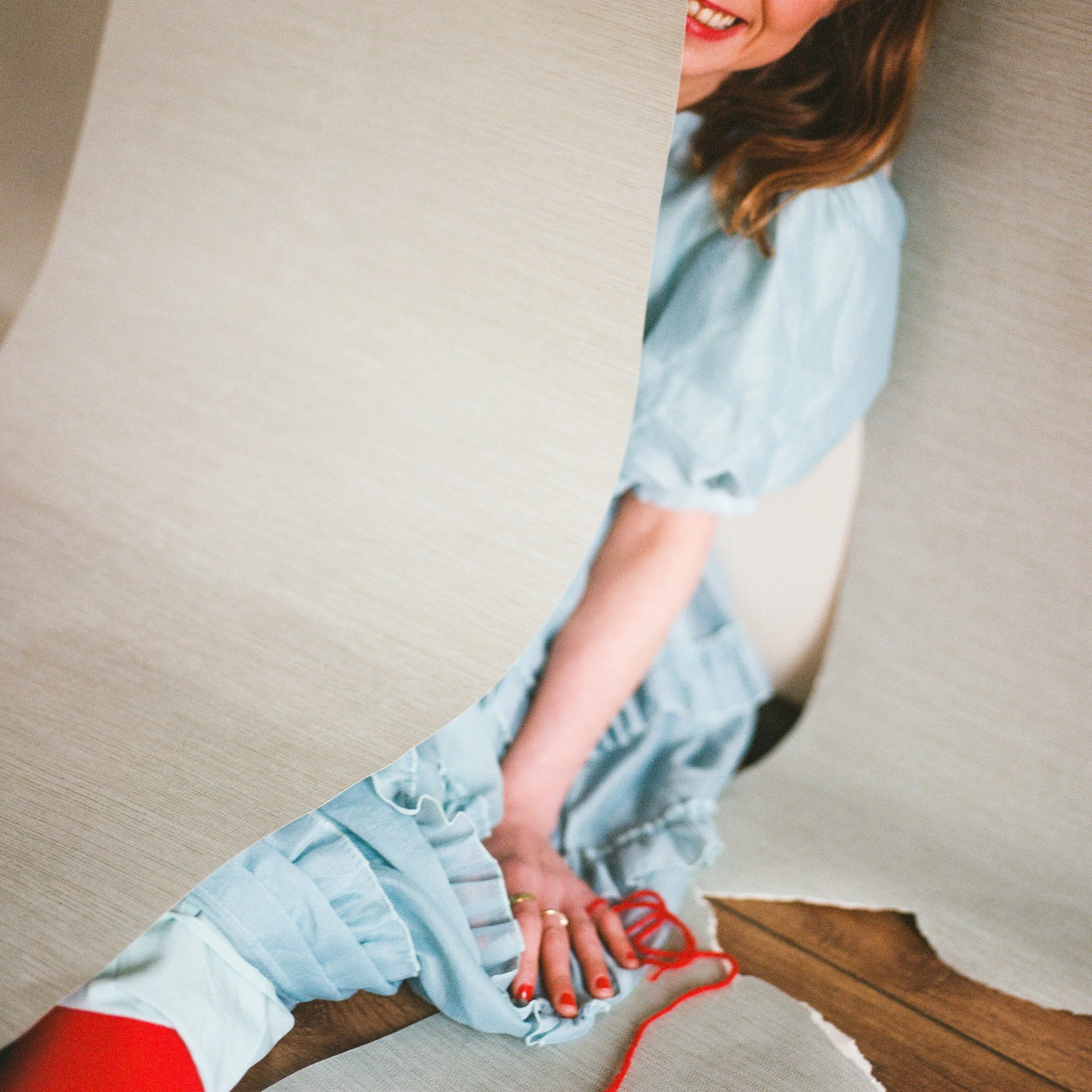 Carla J Easton Album artwork - I Think That I Might Love You A woman in a light blue dress with red shoes and red yarn on her hand, smiling and peeking from behind a beige partition in an indoor setting.