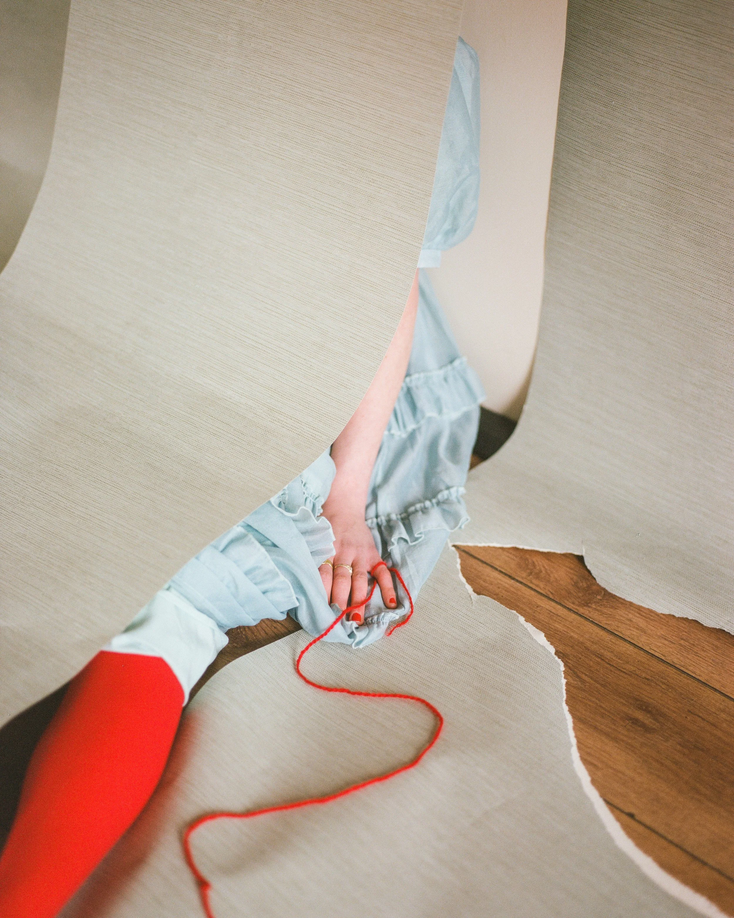 Person in blue dress pulling torn wallpaper from wall, revealing a wooden surface underneath, with a red string in hand.