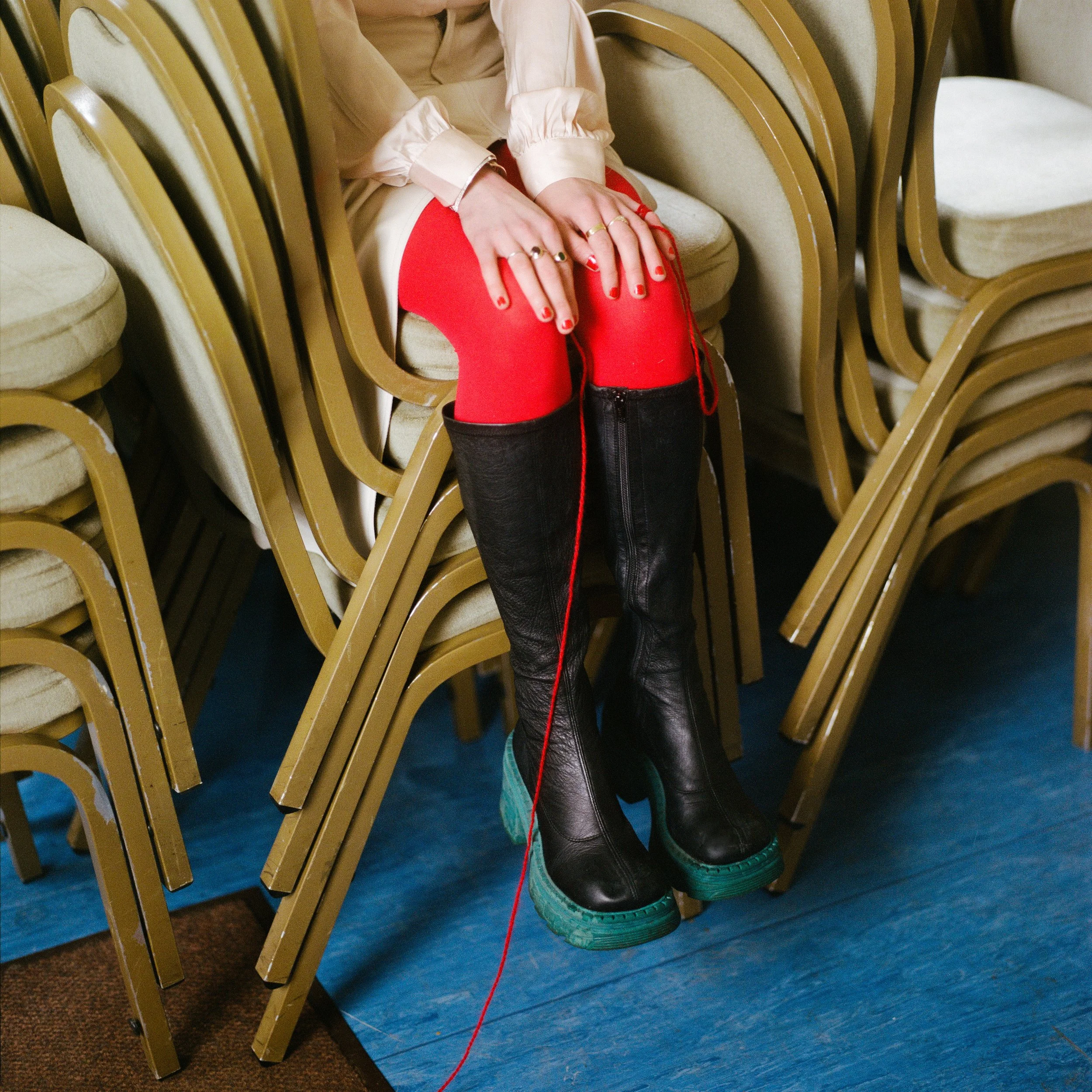 Carla J Easton Album artwork - I Think That I Might Love You A person sitting on stacked chairs with their legs hanging down, wearing red tights and black boots with teal soles, a beige or light pink blouse, and multiple rings on their fingers.