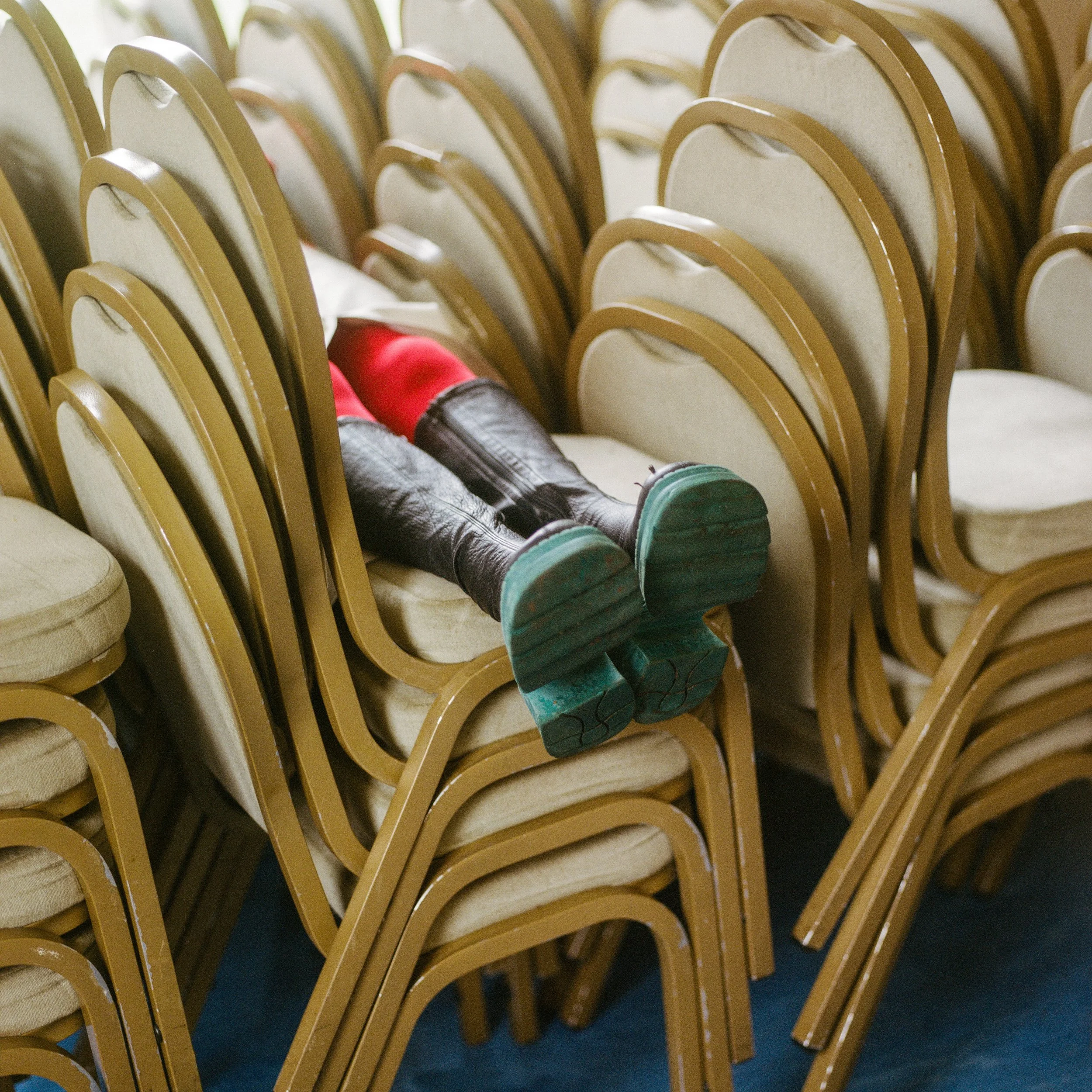Carla J Easton album artwork. Child lying across stacked chairs, legs turned slightly to the side, wearing red shorts, black leather boots, and teal soles.