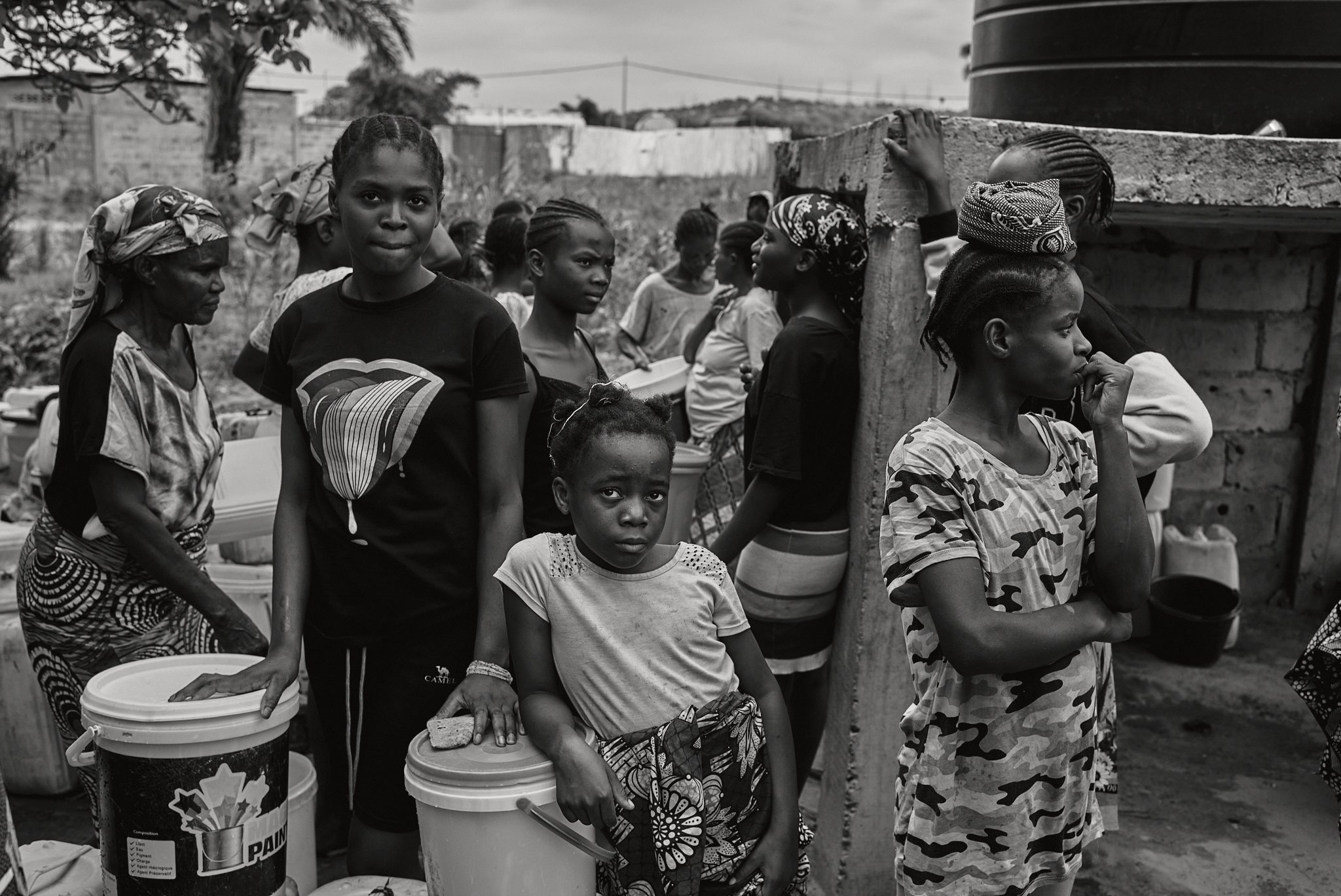 Eine Gruppe von Menschen, überwiegend Frauen und Kinder, stehen in einer Warteschlange im Freien. Es scheint, dass sie auf etwas warten, möglicherweise im Zusammenhang mit Lebensmittelverteilung oder Versorgungsleistungen.