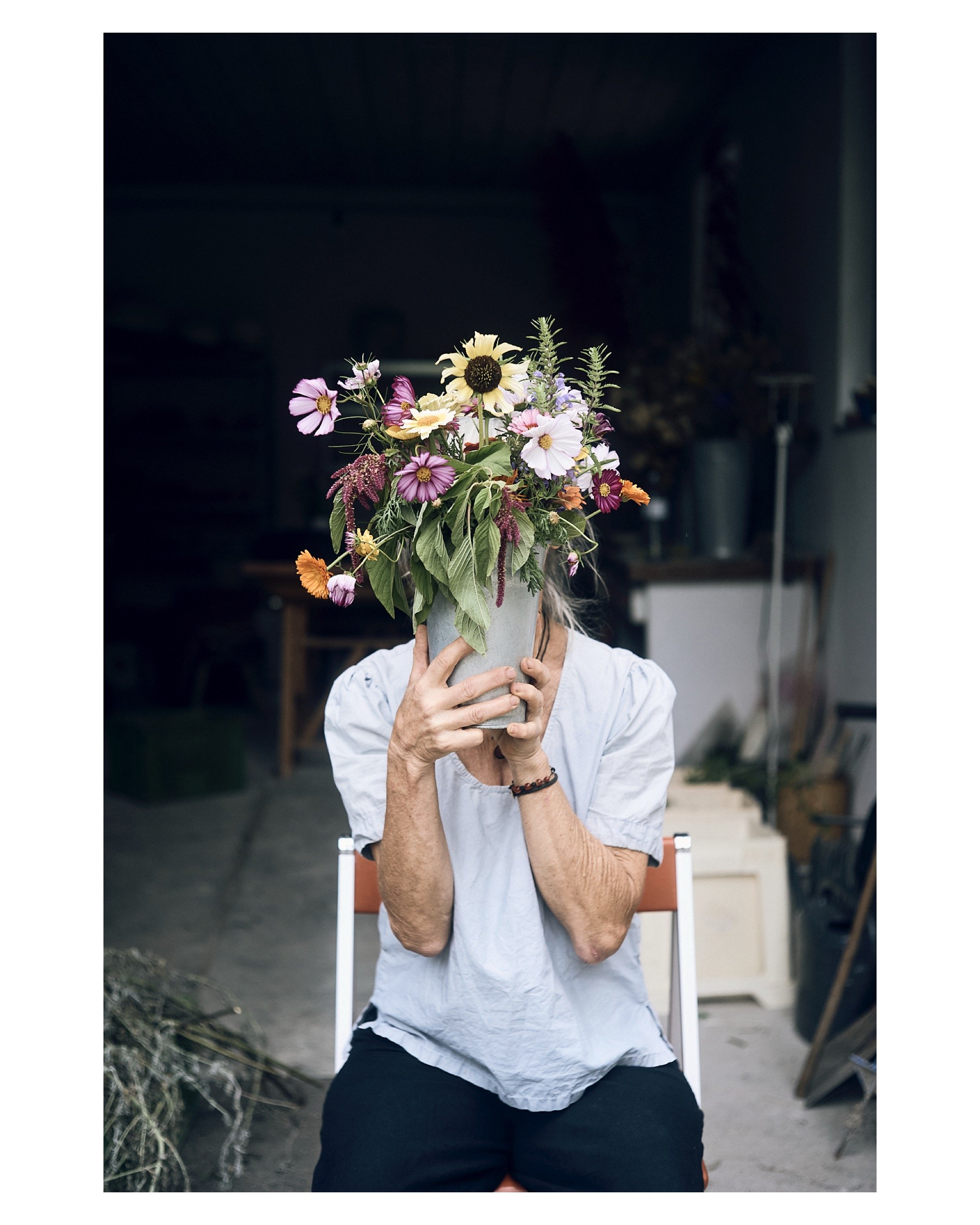 Eine Person sitzt auf einem Stuhl und hält einen Blumenstrauß vor ihr Gesicht, sodass ihr Gesicht nicht sichtbar ist. Der Blumenstrauß besteht aus verschiedenen bunten Blumen und Grünzeug.