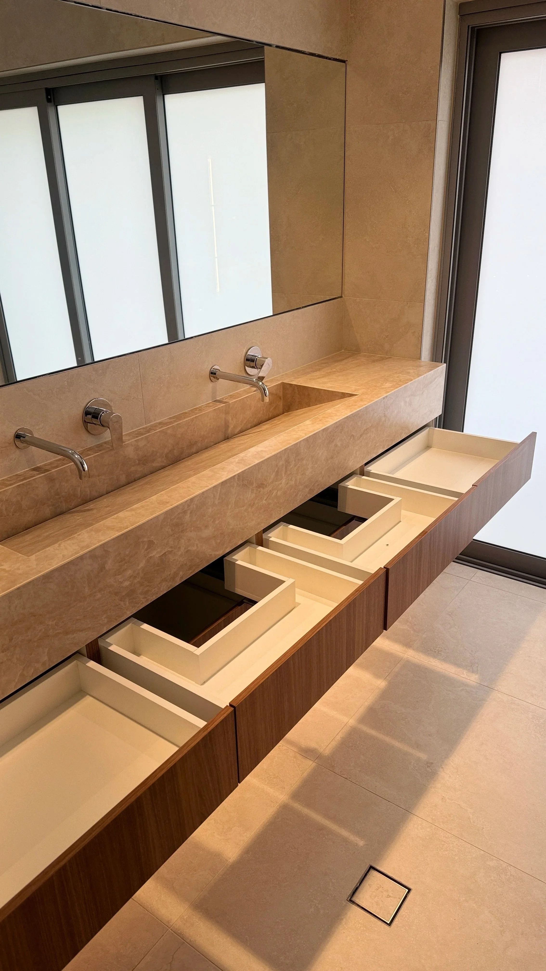 Modern bathroom with a beige marble sink countertop, three silver faucets, a large mirror, and open cabinets below the counter, adjacent to a frosted glass door.