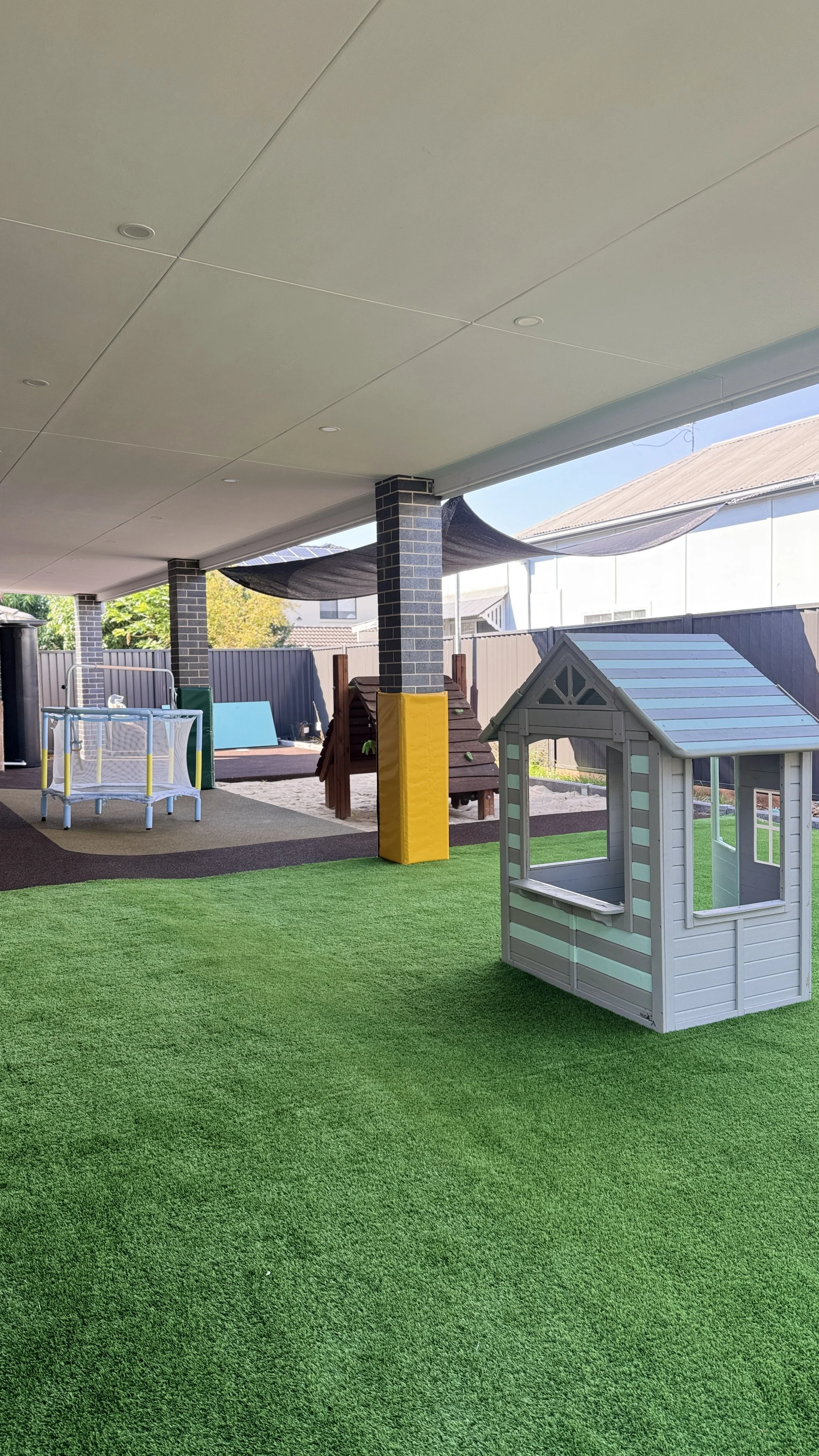 Play area with artificial green grass, small white playhouse, and wooden climbing structure under a covered patio, with outdoor setting in the background.