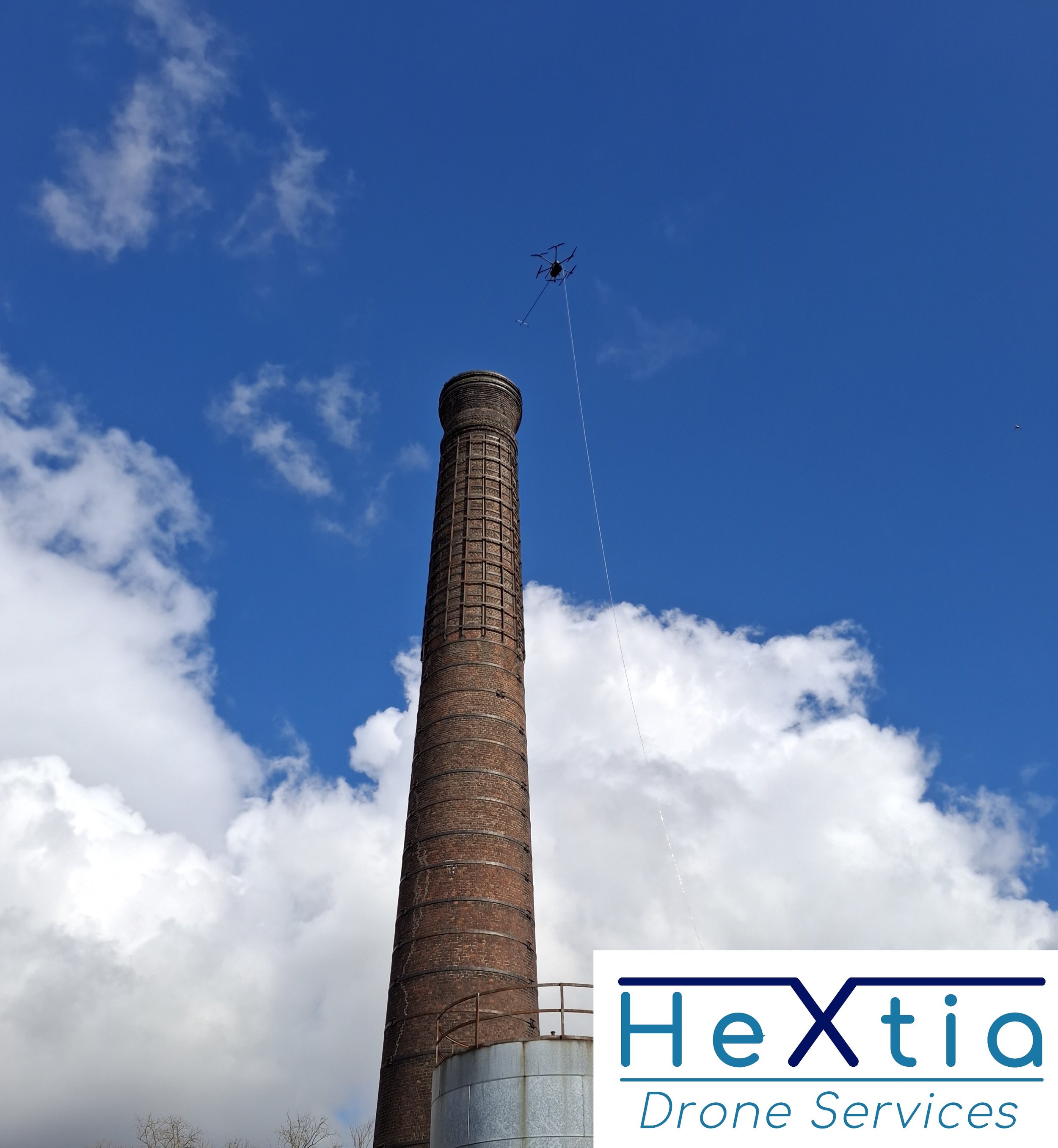 Une cheminée en brique de grande taille avec un drone volant au-dessus, dans un ciel bleu avec quelques nuages, et un logo en bas à droite pour HexTia Drone Services.