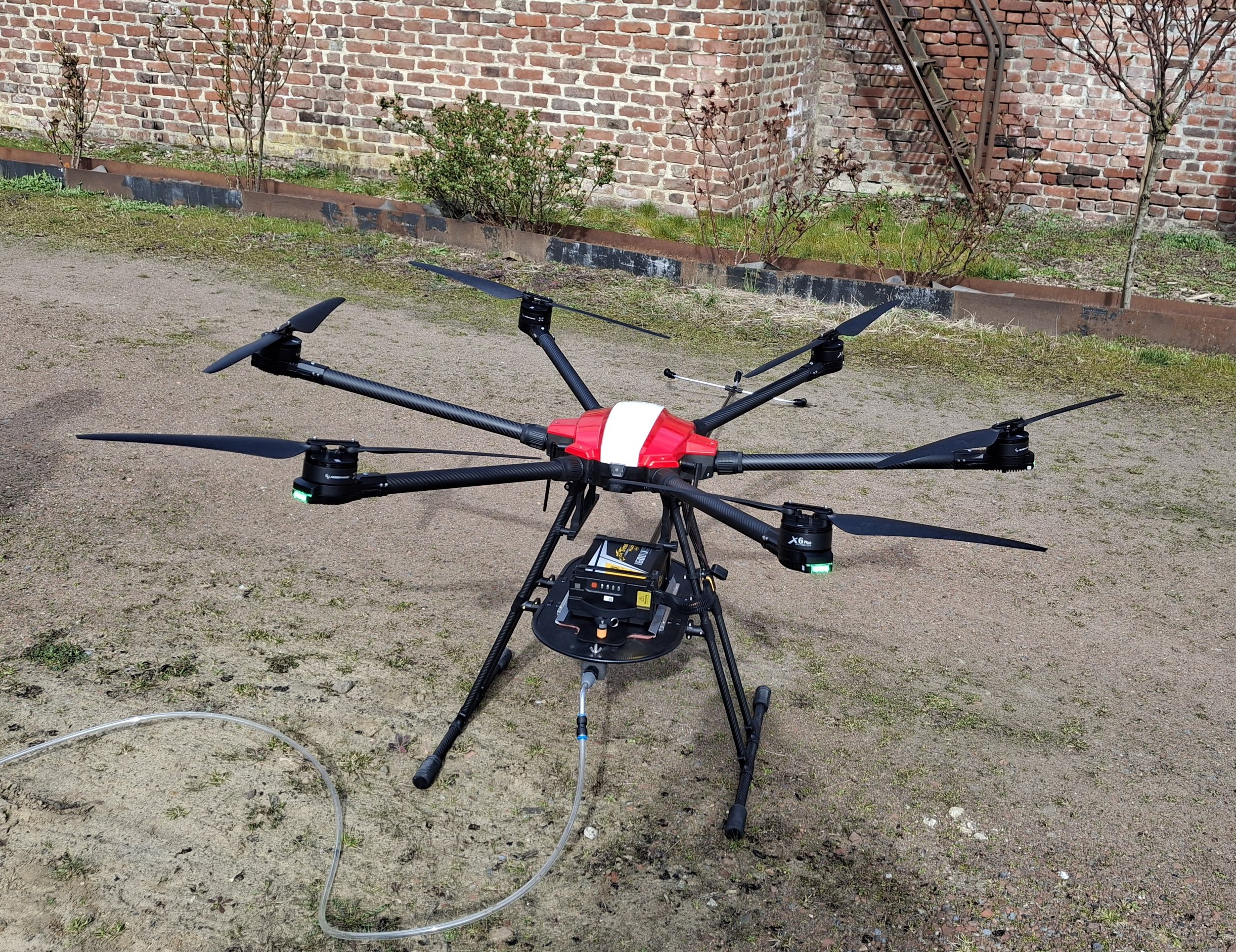 Un drone multirotor posé sur le sol en extérieur, près d'une paroi en briques, avec des jeunes arbres en arrière-plan.