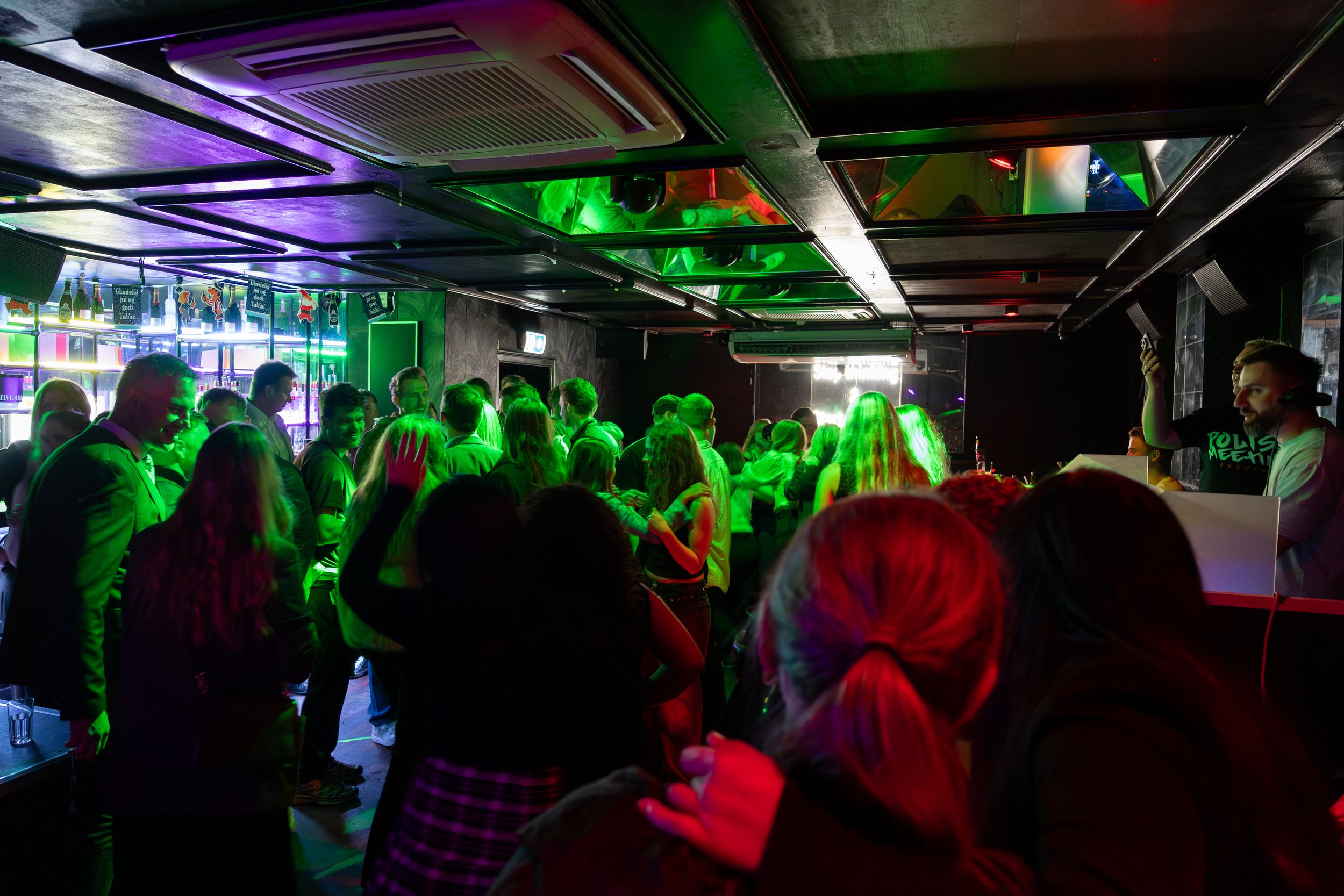 Crowd of people dancing at a nightclub with colorful green, purple, and red lighting and DJ playing.
