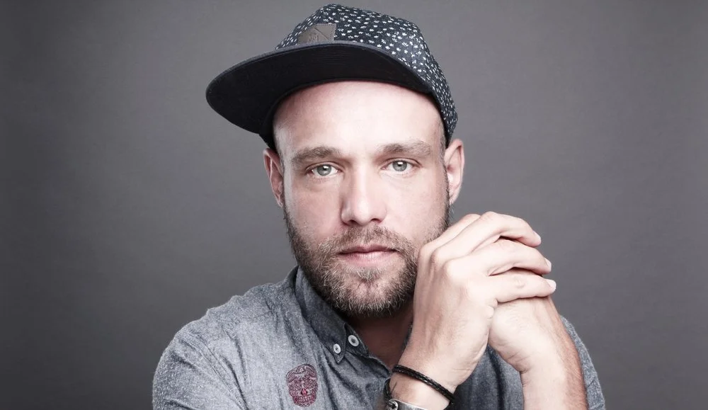 A man with a beard and mustache wearing a gray shirt and a black cap with white patterns, posing against a gray background.