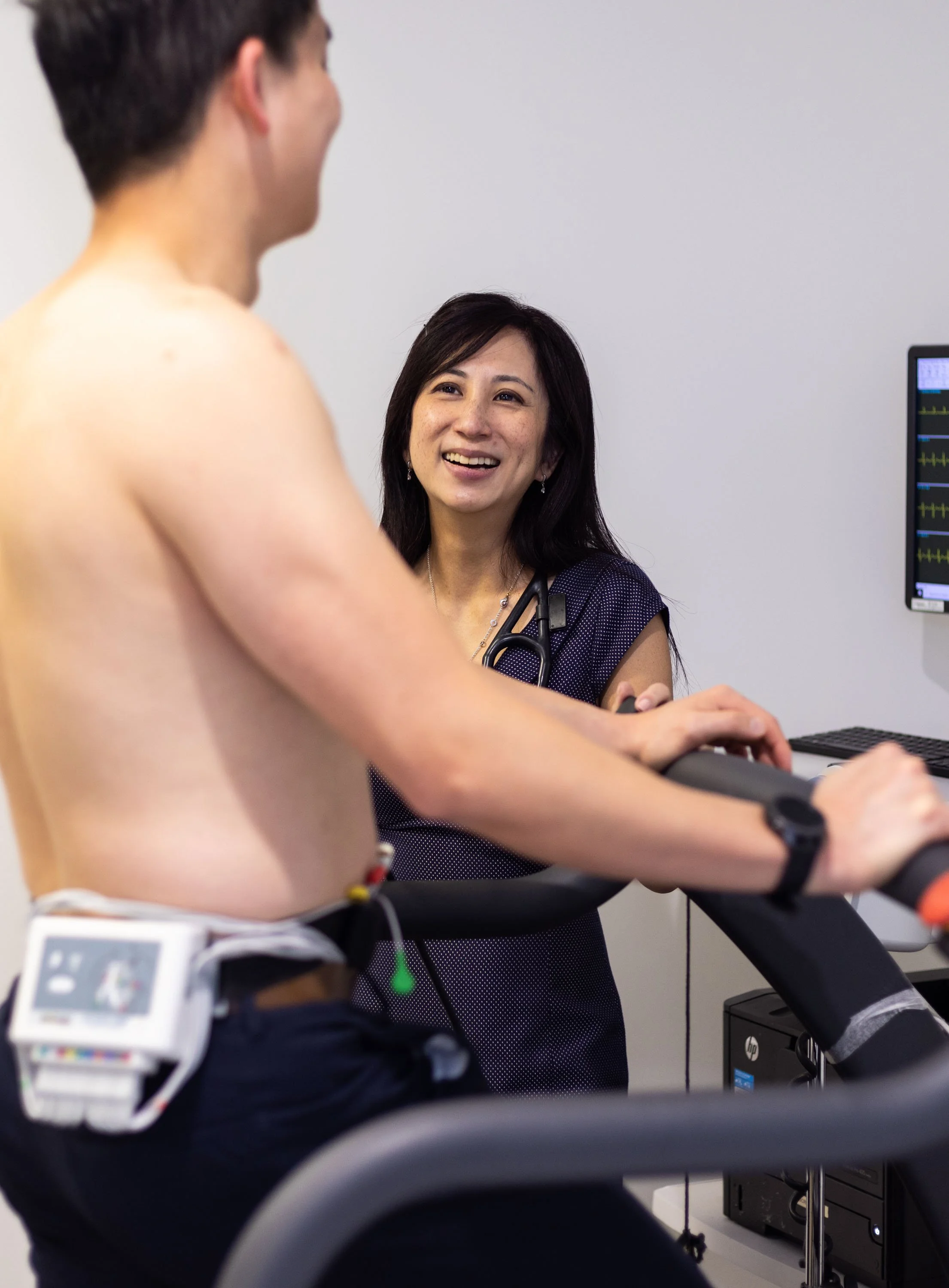 A healthcare professional talking to a young man on a treadmill during a medical examination, with a monitor displaying vital signs in the background.