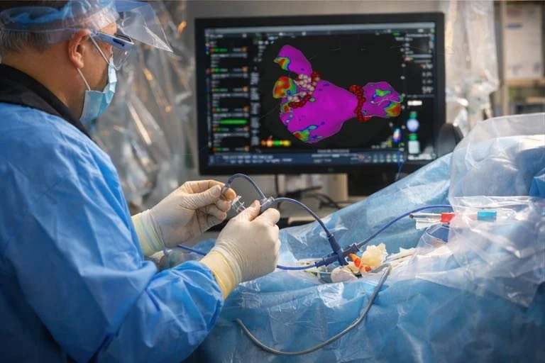 An electrophysiologist in scrubs performing an ablation procedure in a catheter lab, with a computer monitor displaying a colourful 3D model.