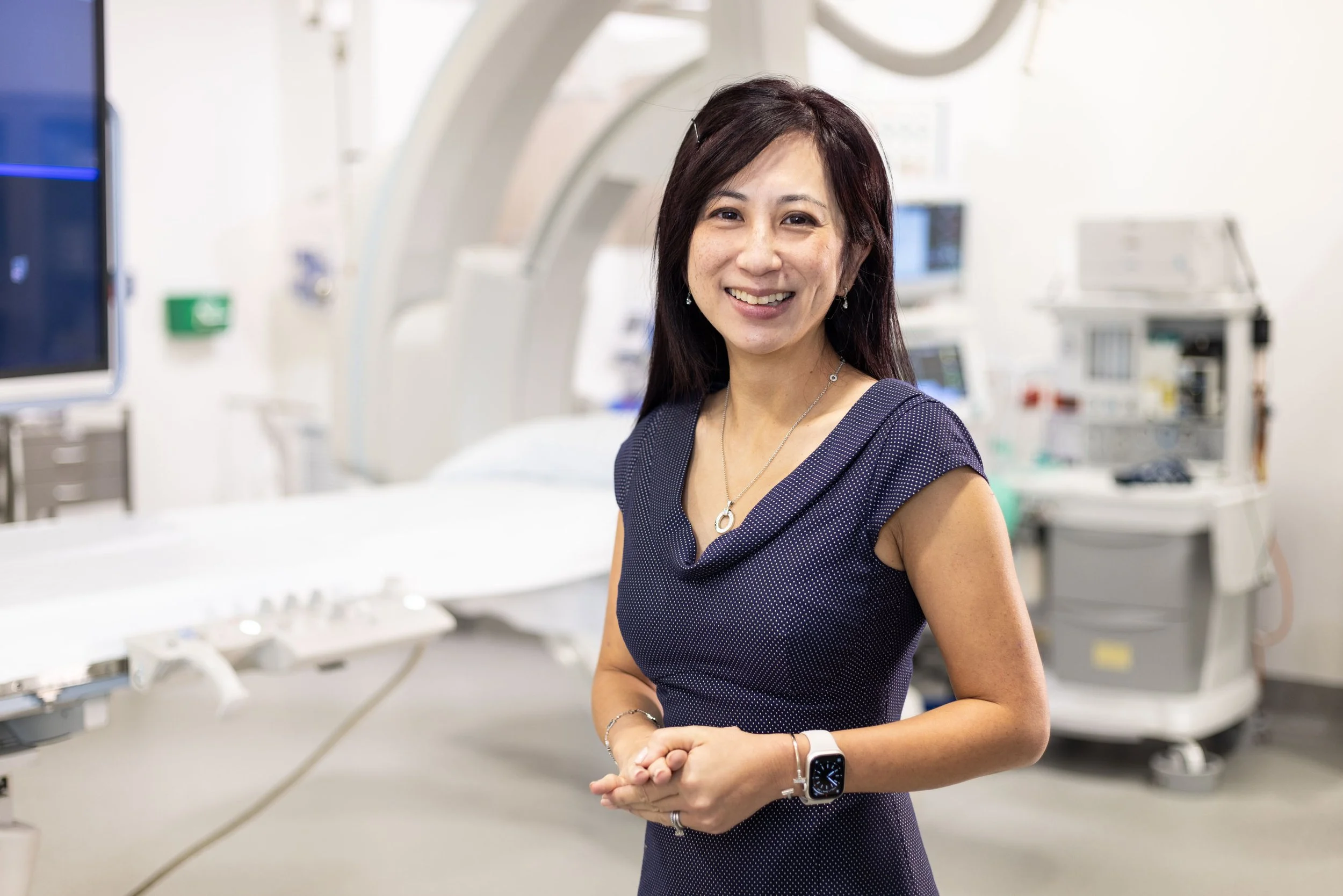 A woman in a navy blue dress with short sleeves, smiles at the camera, standing in a medical or hospital room with medical equipment in the background.