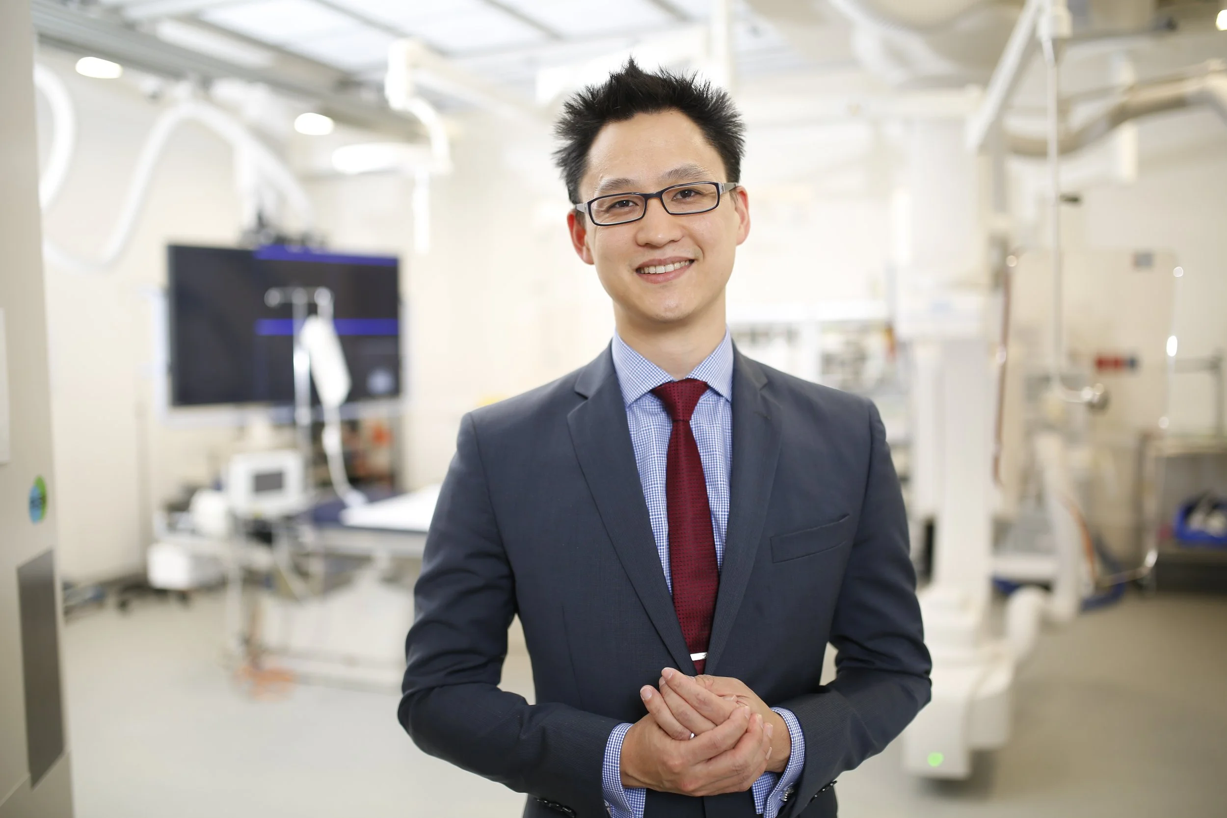 Man in business suit and red tie smiling in a laboratory with medical equipment and monitors in the background.