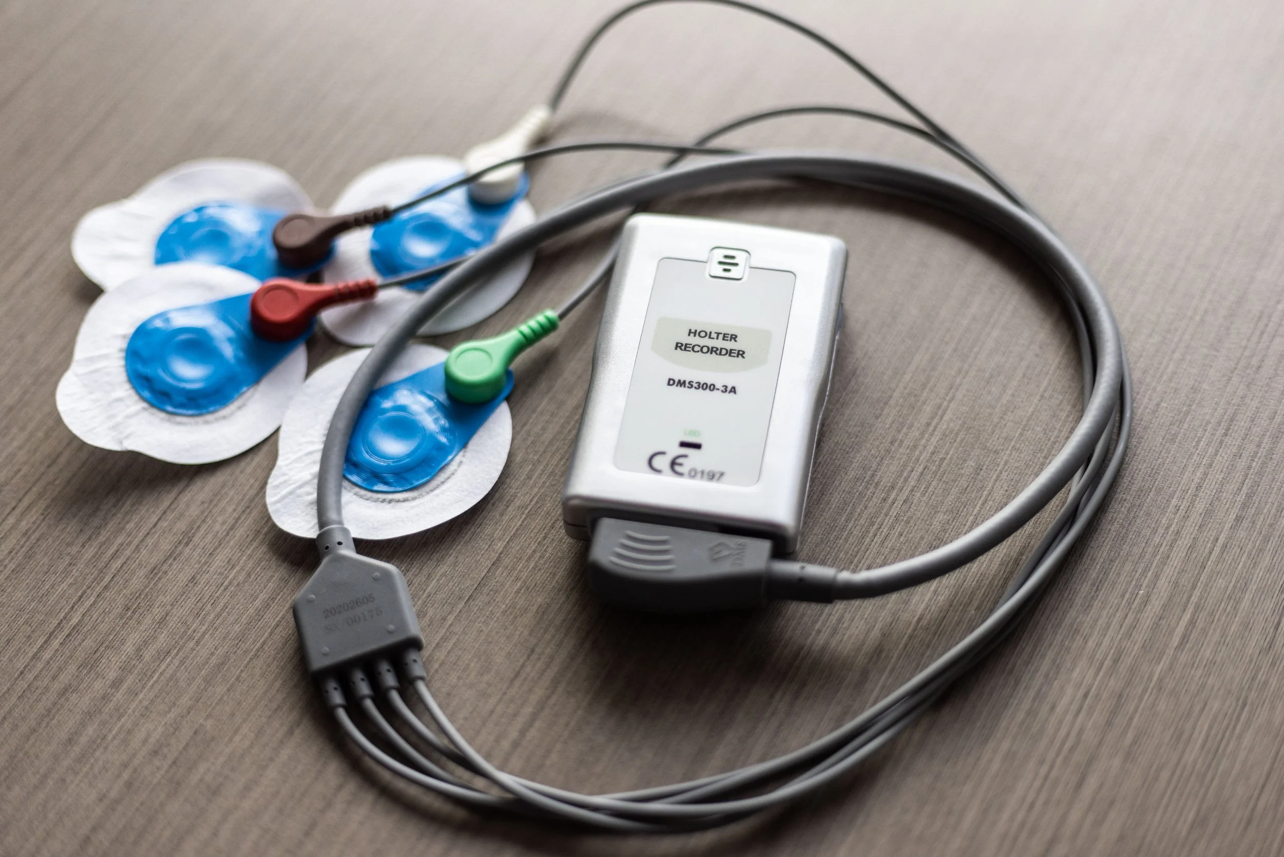 A Holter monitor device connected to three electrode patches with colored wires, placed on a wooden surface.