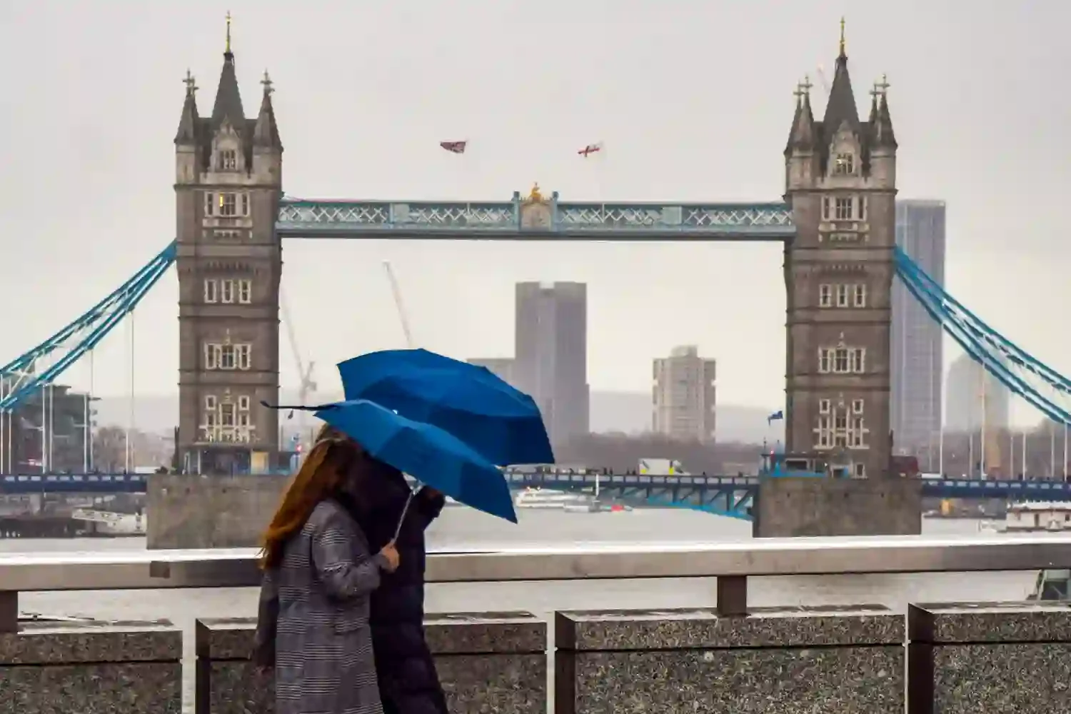 To personer som går sammen under blå paraplyer foran Tower Bridge i London, med regn og bybygg i bakgrunnen.