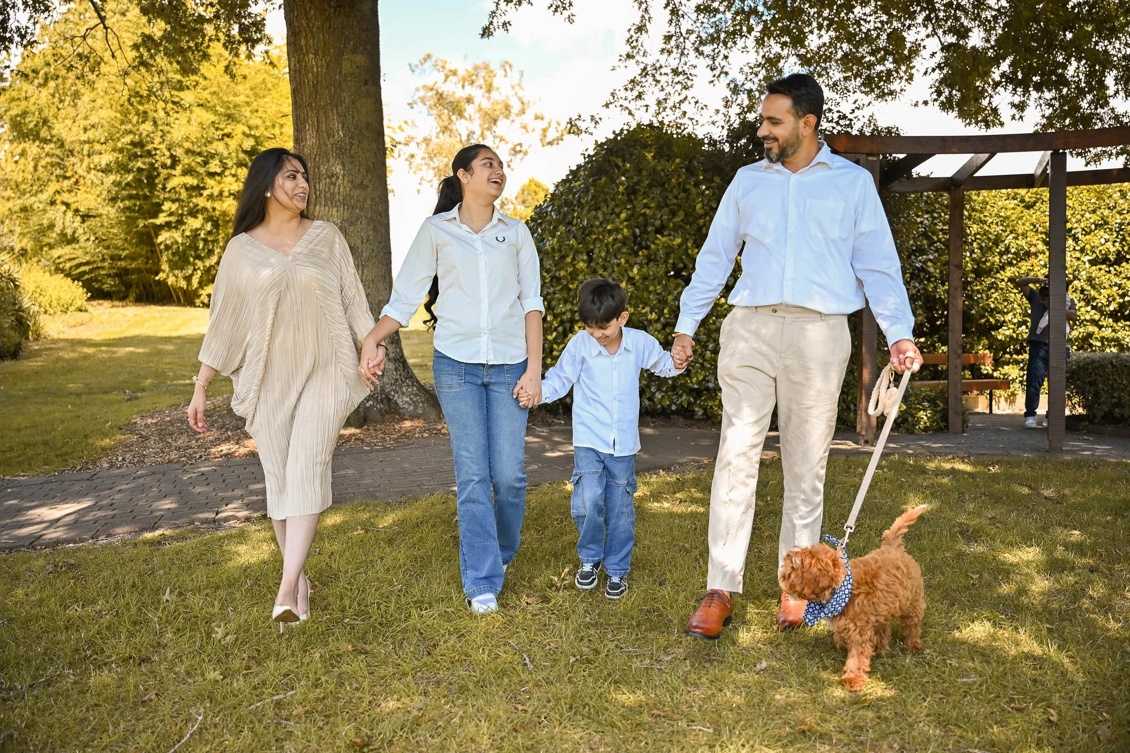 A family of five, including a dog, walking hand in hand outdoors in a park on a sunny day.
