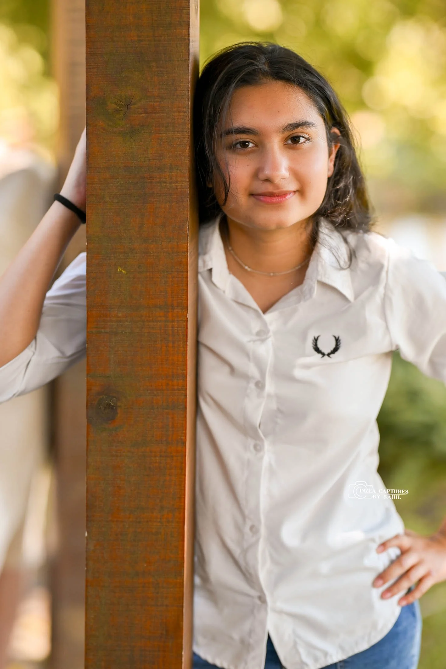A young woman with shoulder-length dark hair, wearing a white blouse with a black logo on it, stands outdoors next to a wooden post. She has one hand on her hip and the other hand resting on the post, with a slight smile and a background of blurred g