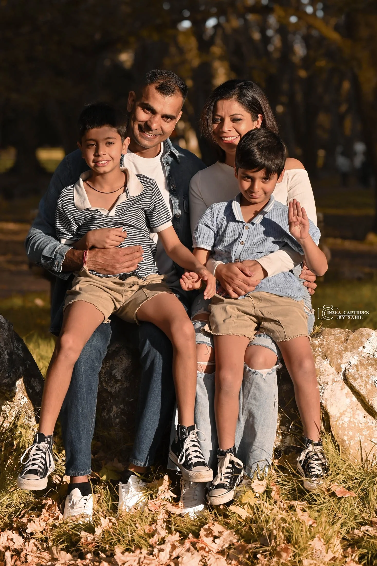 Family of four sitting on rocks outdoors in autumn, smiling at camera.