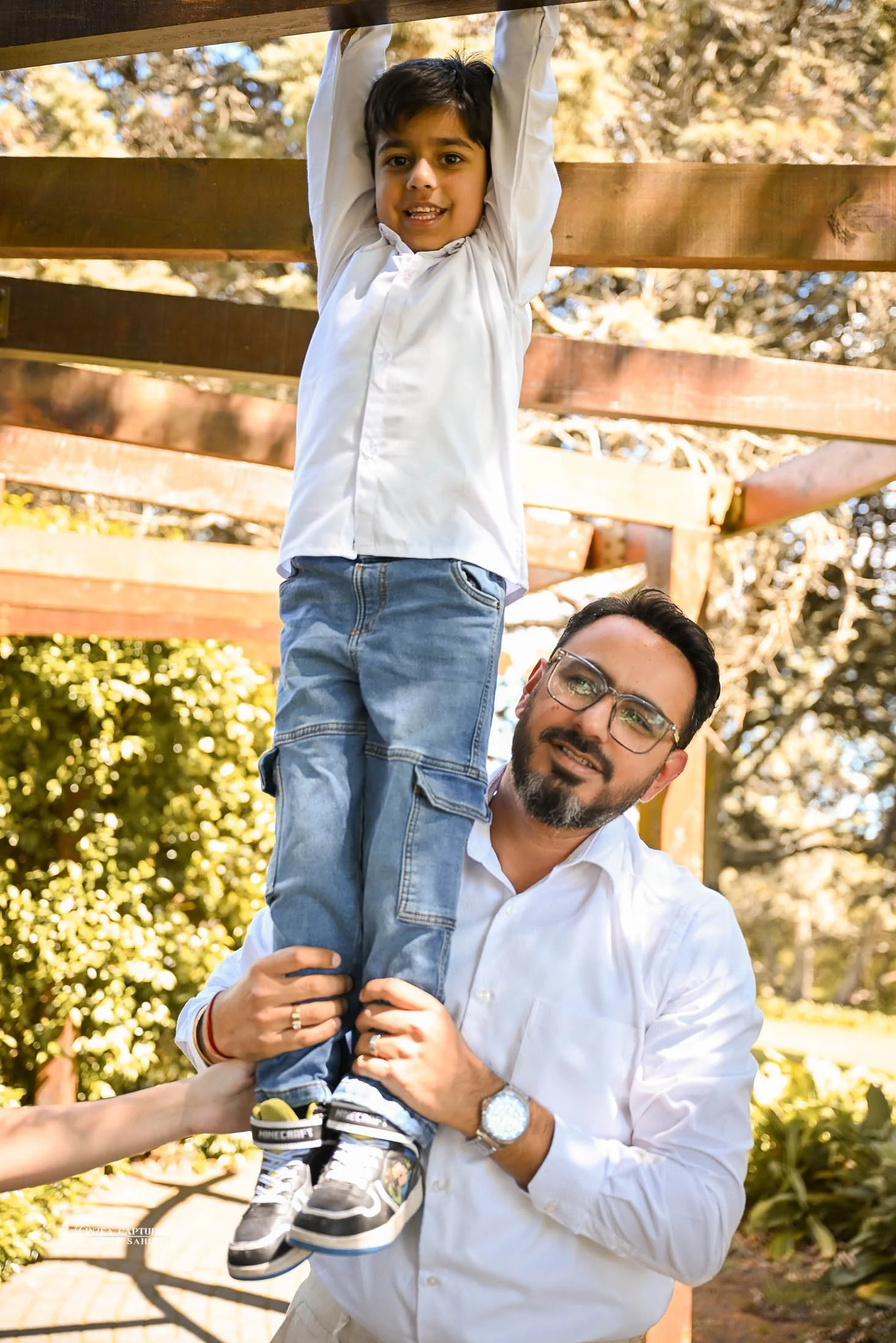 A man wearing glasses and a white shirt holding up a young boy with a white shirt and blue jeans on his shoulders outdoors.