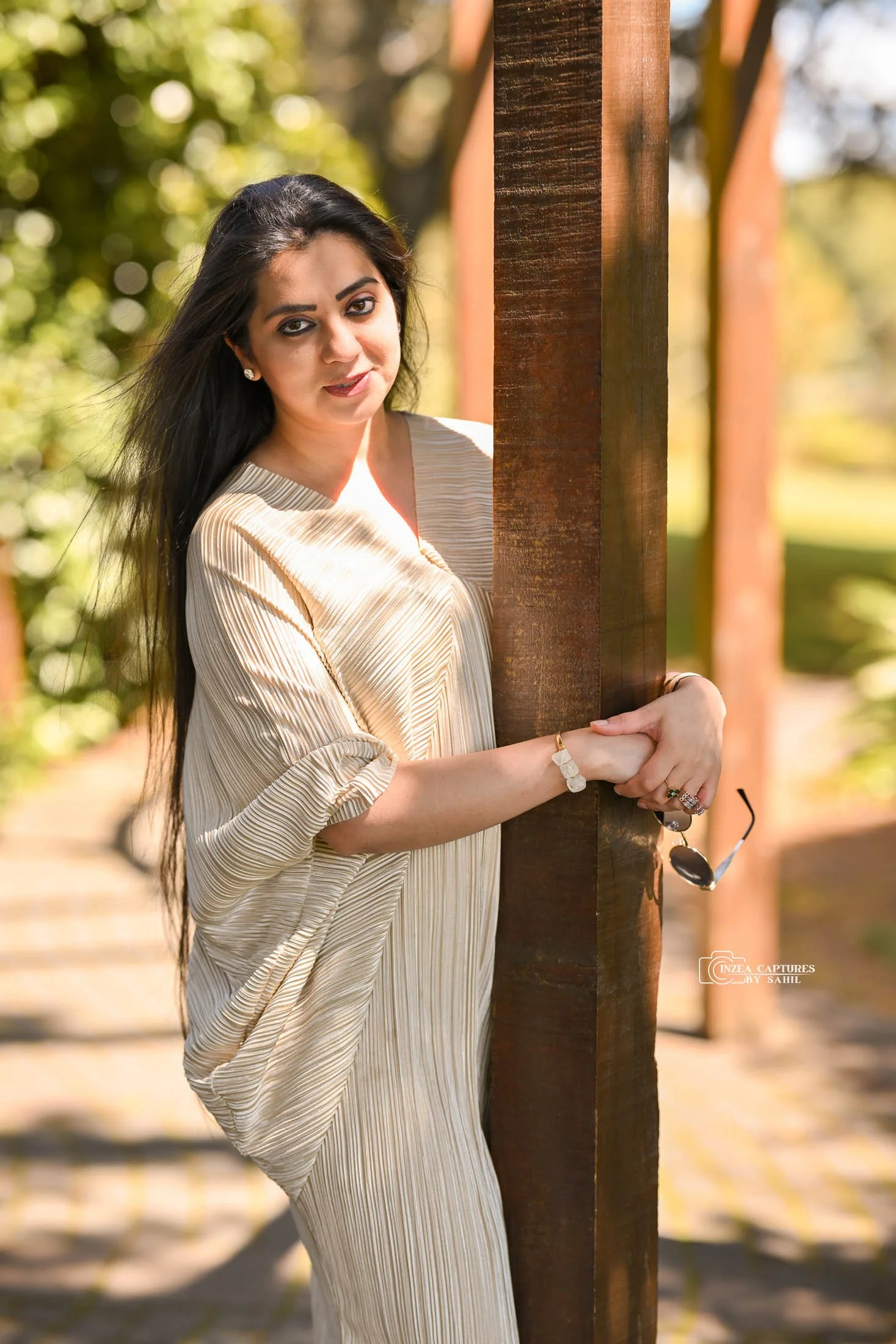 A woman with long black hair and earrings is holding onto a wooden post outdoors on a sunny day, with trees and blurred greenery in the background.