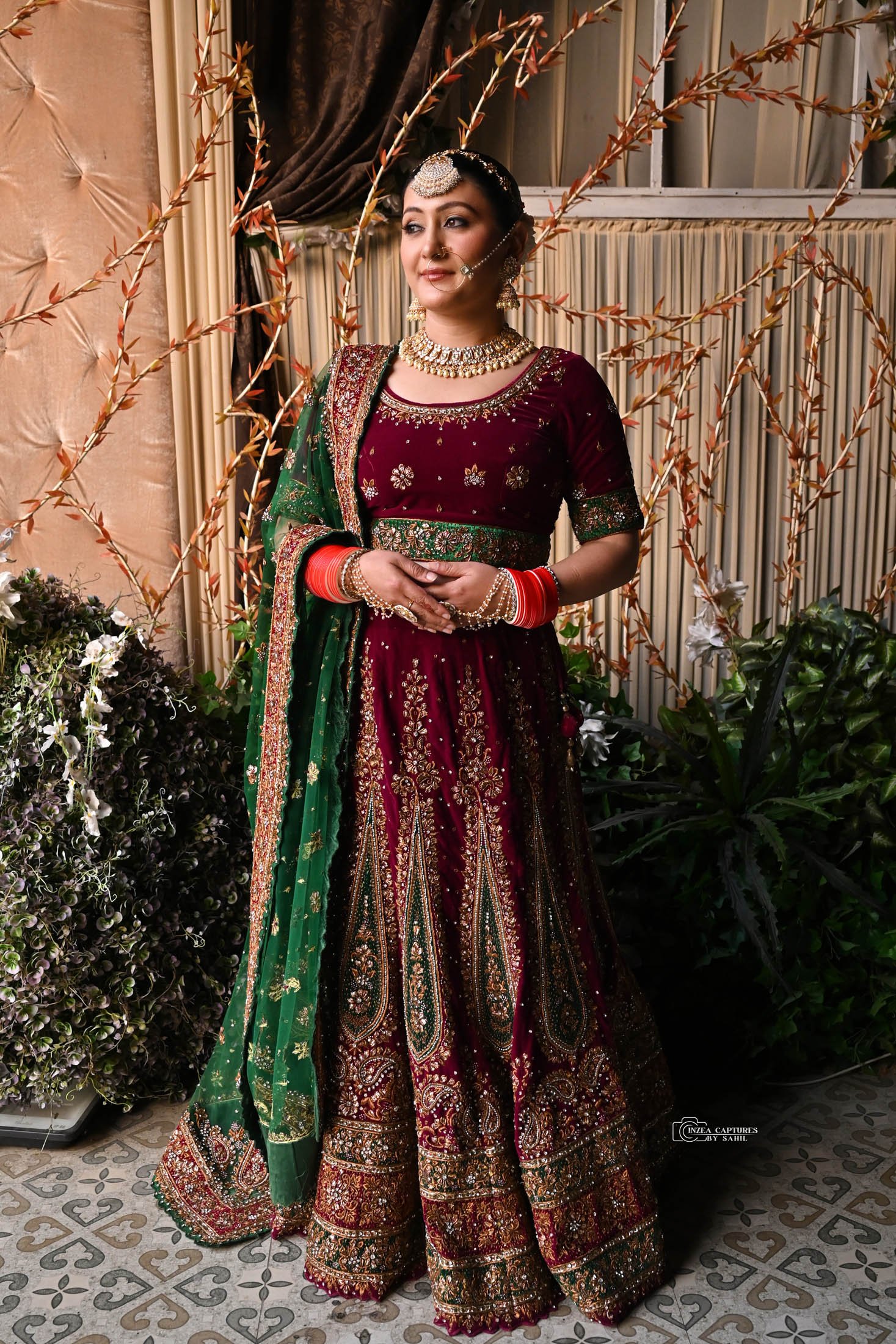 A woman in an ornate maroon and green traditional Indian bridal dress with gold embroidery, standing indoors against a backdrop with plants and a window.