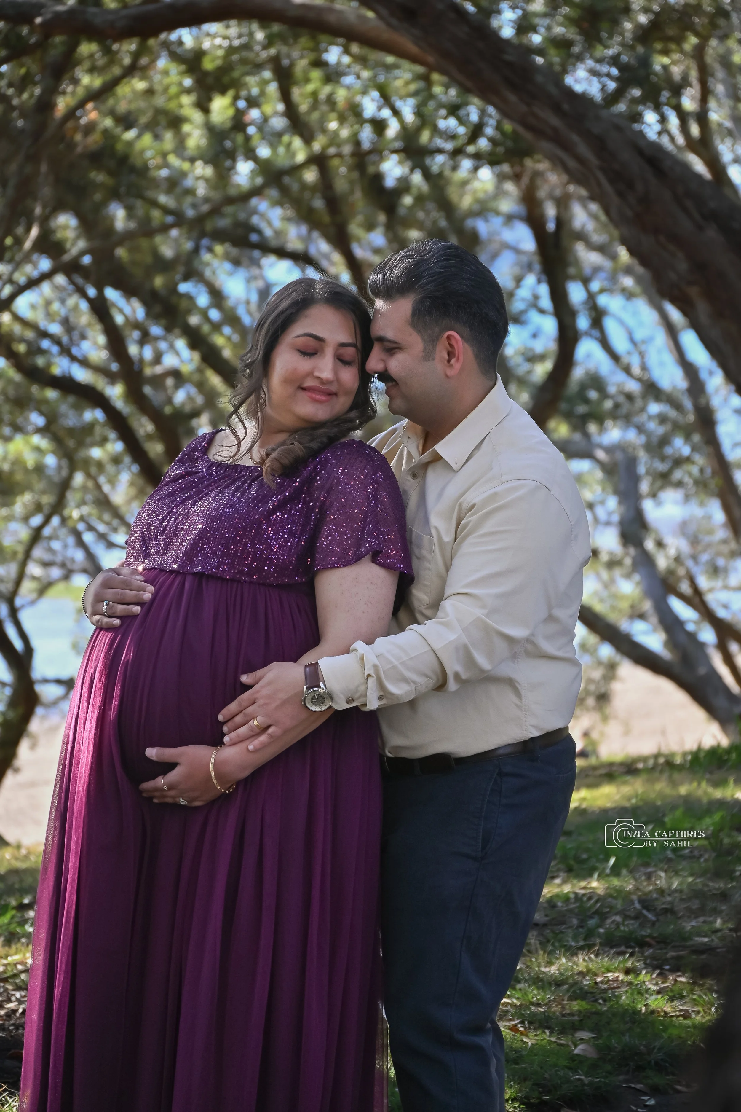Pregnant woman in a purple dress with her partner, both smiling and touching her belly, outdoors under a large tree with leaves and blue sky visible in the background.