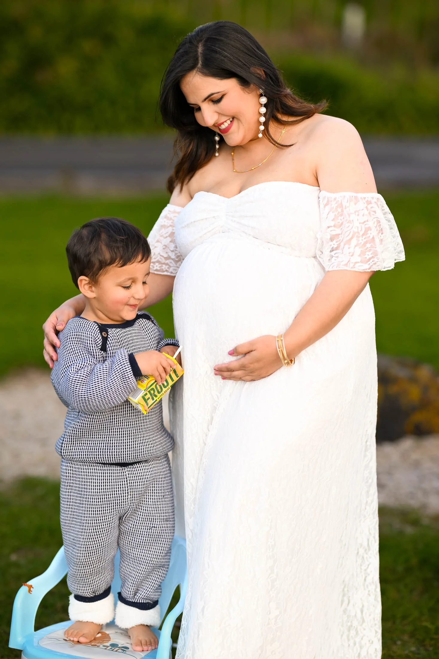 A pregnant woman dressed in a white off-shoulder dress smiling and looking down at a young boy who is holding a juice box, standing on a small chair outdoors in a grassy area.