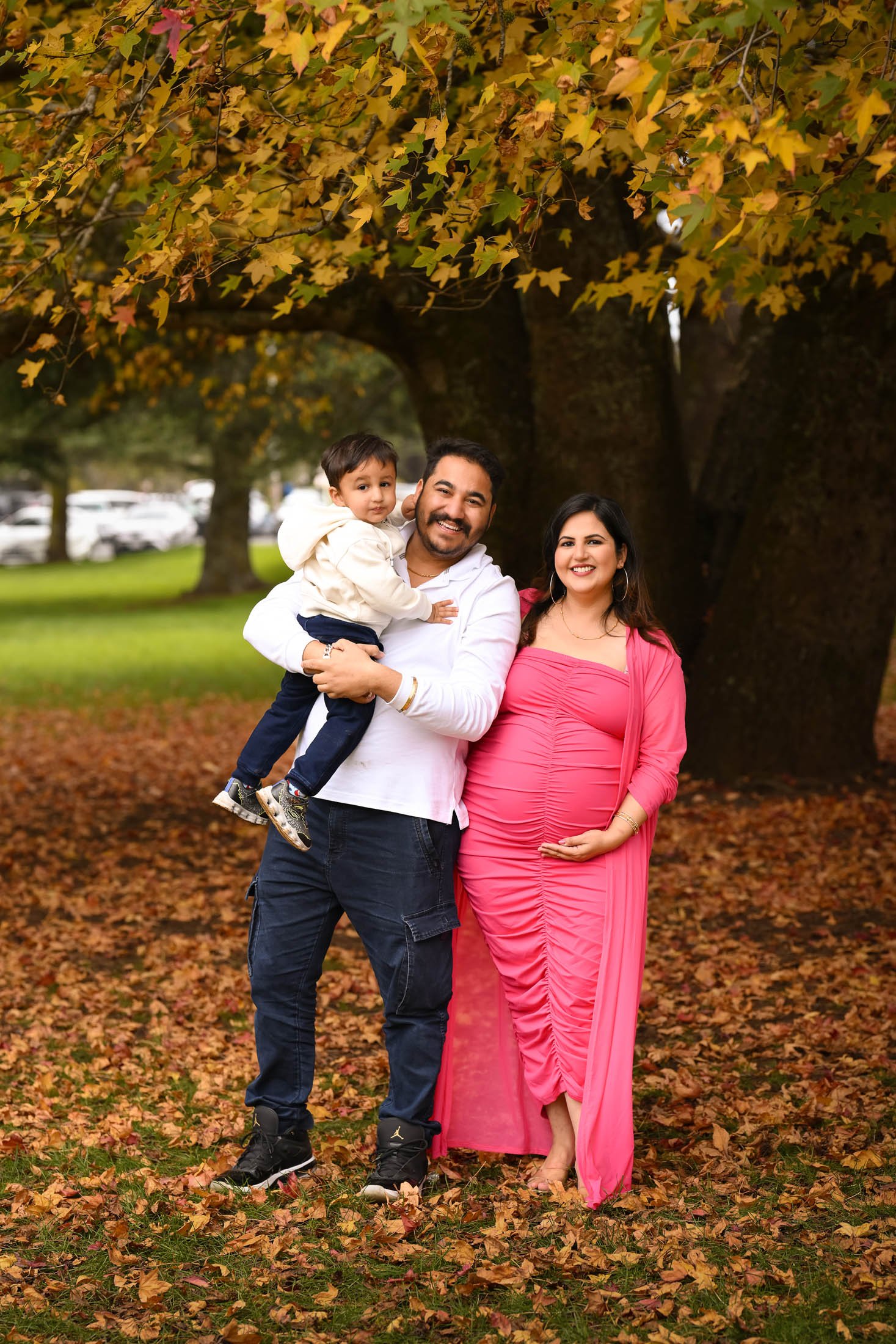 A family of three standing outdoors under a large tree with yellow and green leaves during autumn. The father is holding a young boy, and the mother, who is pregnant, is standing beside them. All are smiling, and the ground is covered with fallen lea
