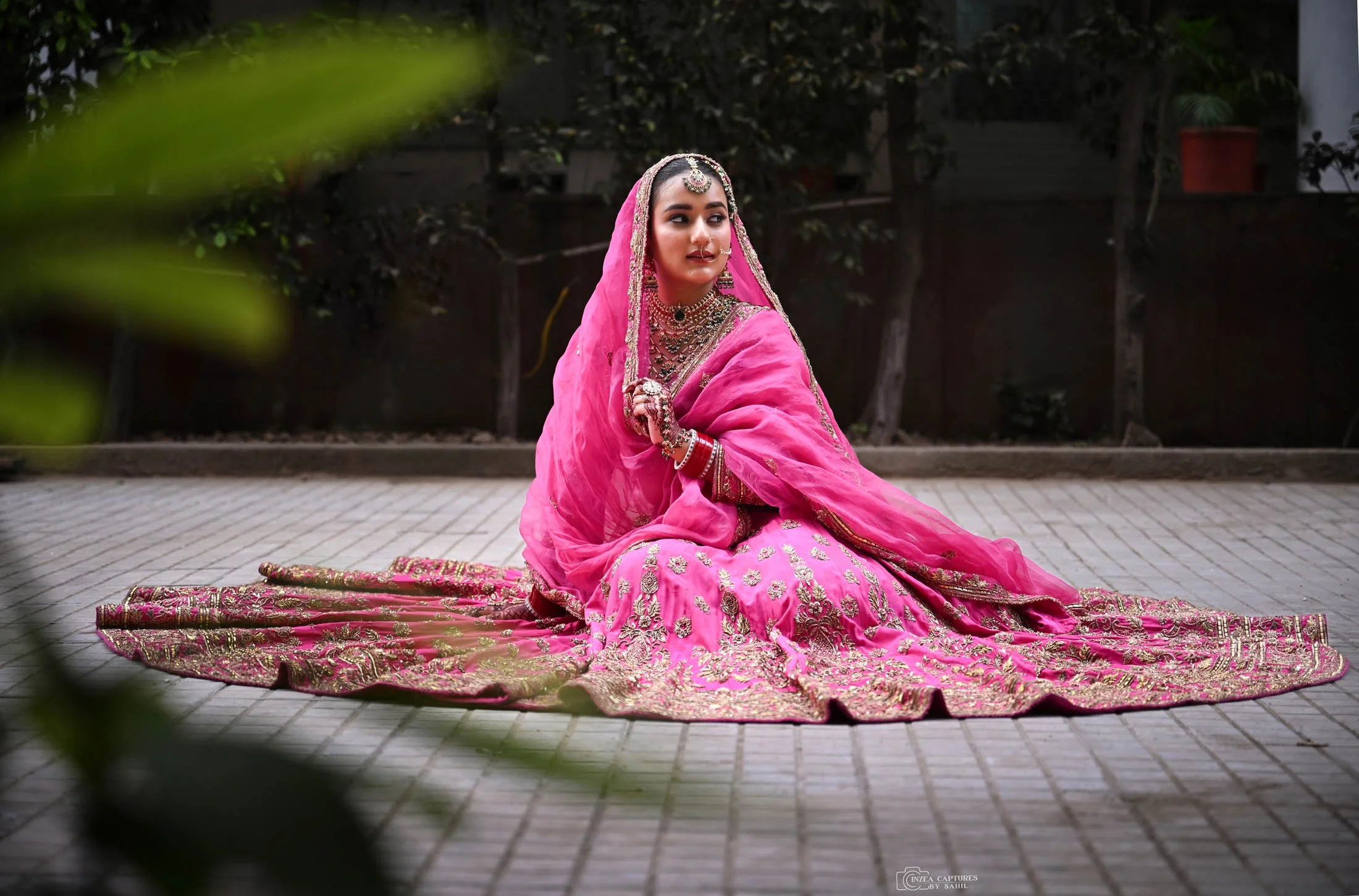 A woman dressed in a traditional pink Indian bridal saree with intricate gold embroidery, sitting on a paved outdoor area, adorned with jewelry including necklaces, earrings, and bangles.