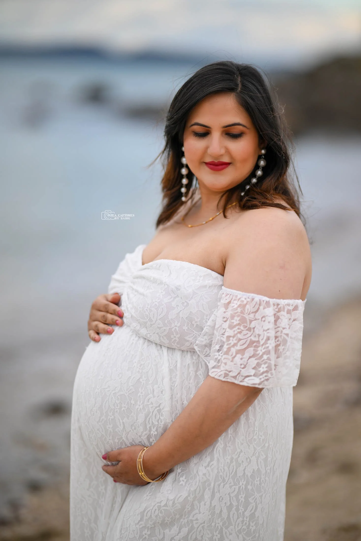 Pregnant woman in a white lace off-shoulder dress standing on a beach, gently cradling her baby bump with her eyes closed and a peaceful smile.
